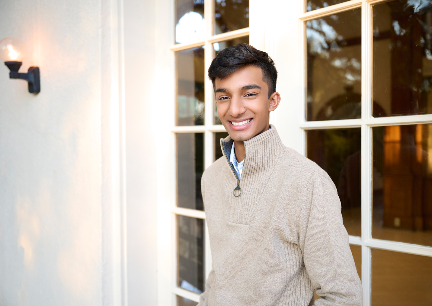 Young man smiling outside a house near a white-framed window, wearing a beige sweater.
