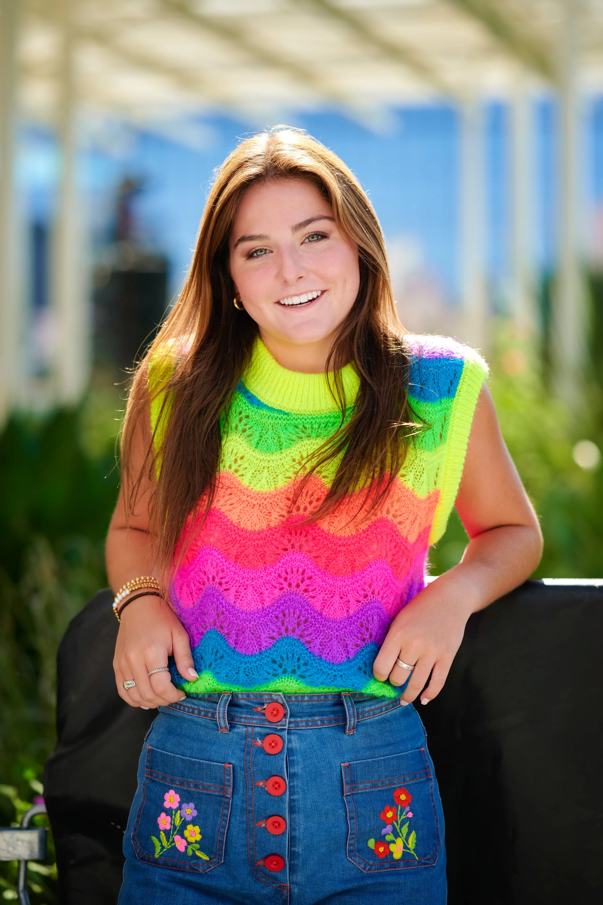 A young woman wearing a bright rainbow-colored knit tank top and a denim skirt with colorful floral embroidery, smiling outdoors on a sunny day.