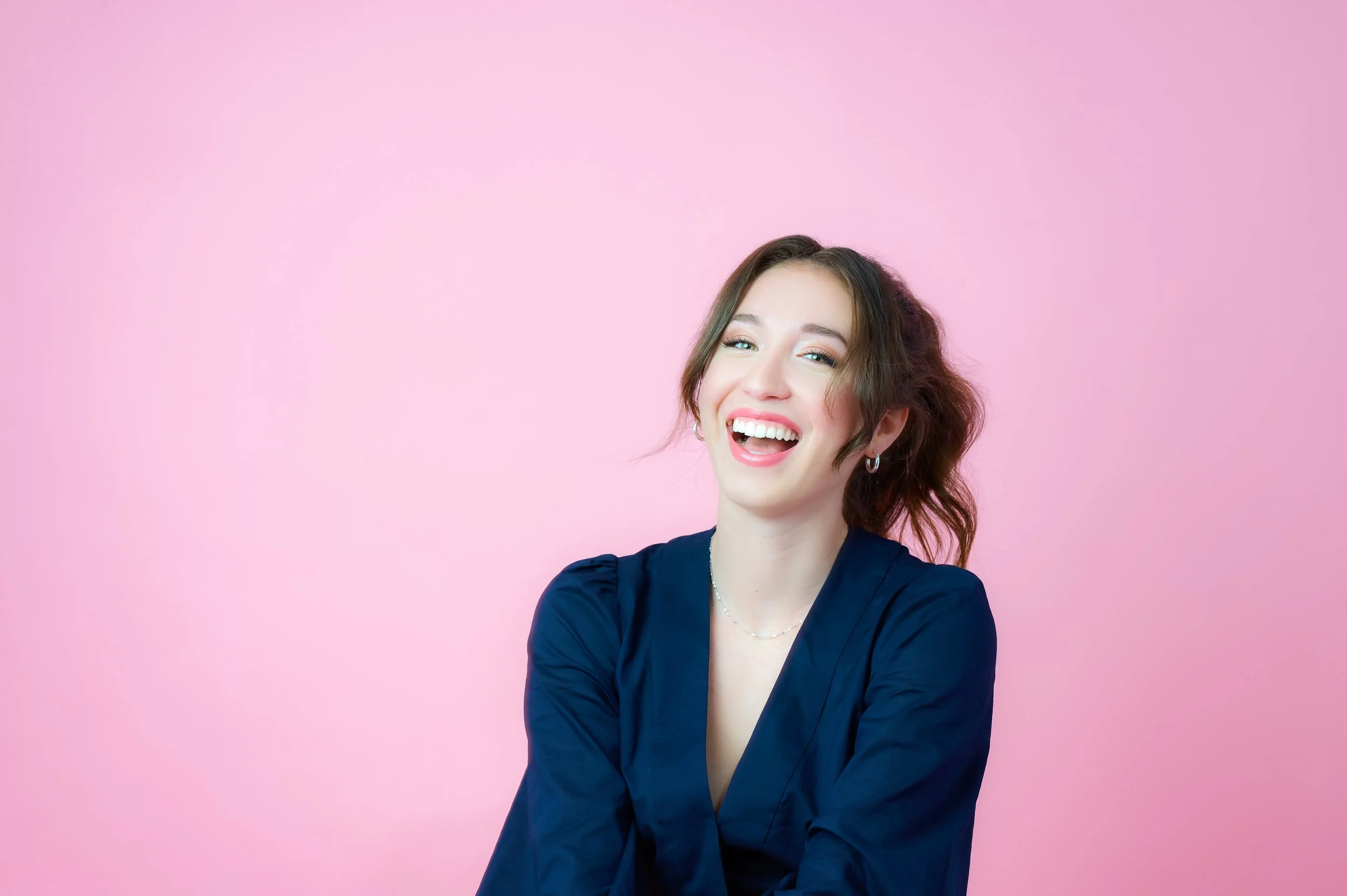 A woman with shoulder-length brown hair, smiling and wearing a dark blue blouse, against a pink background.