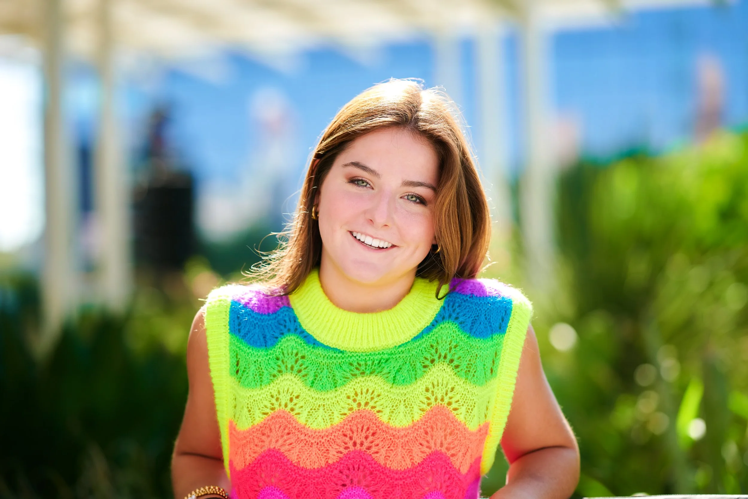 A young woman with shoulder-length brown hair, wearing a brightly colored, sleeveless, rainbow-patterned crocheted top, smiling outdoors with greenery and blue structures in the background.