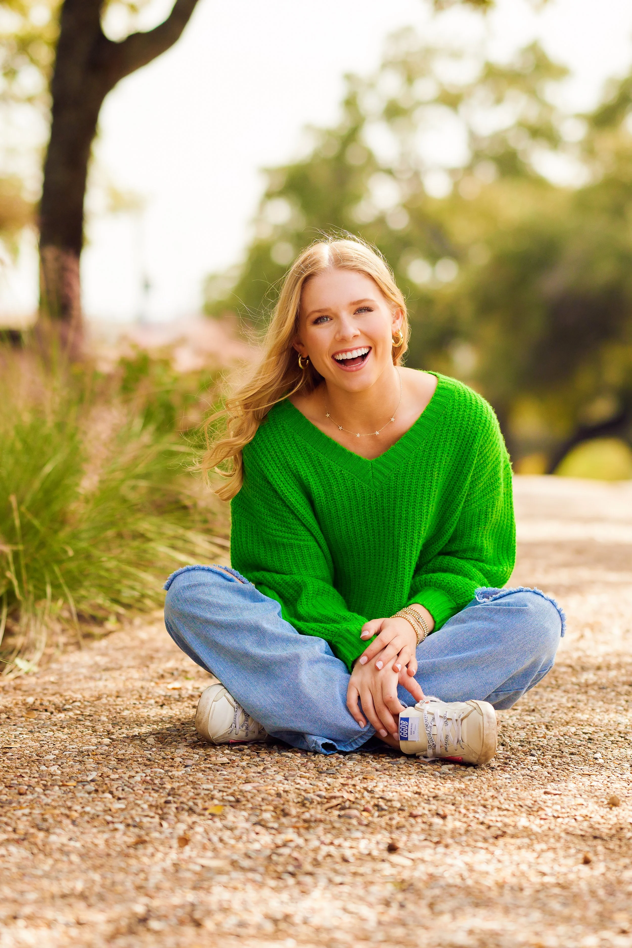 A young woman with blonde hair sitting cross-legged on a gravel path, smiling, wearing a bright green sweater, ripped blue jeans, and white sneakers in an outdoor setting with trees and greenery in the background.