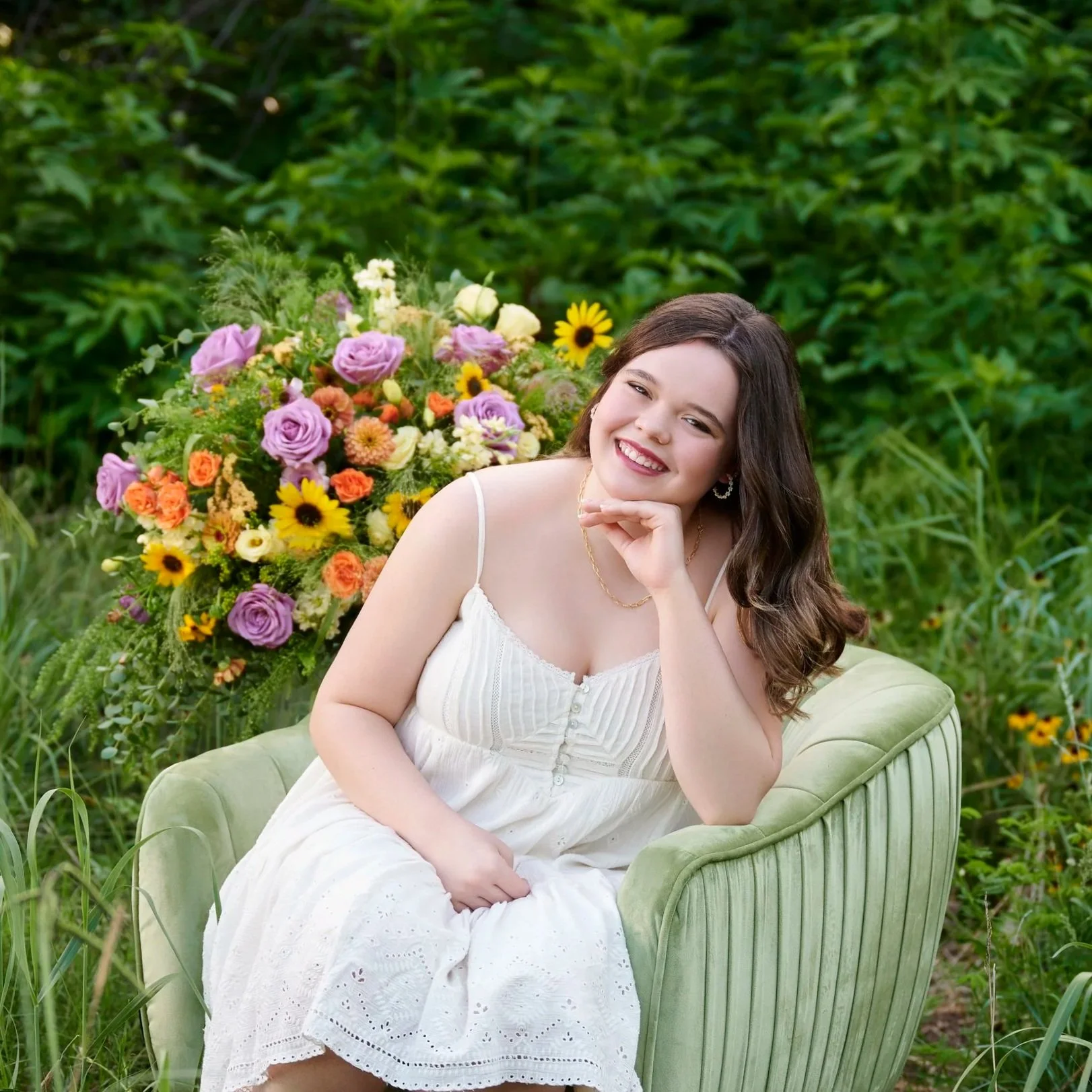 A young woman in a white sundress sitting on a green velvet armchair outdoors, smiling, with a large colorful flower arrangement in the background.