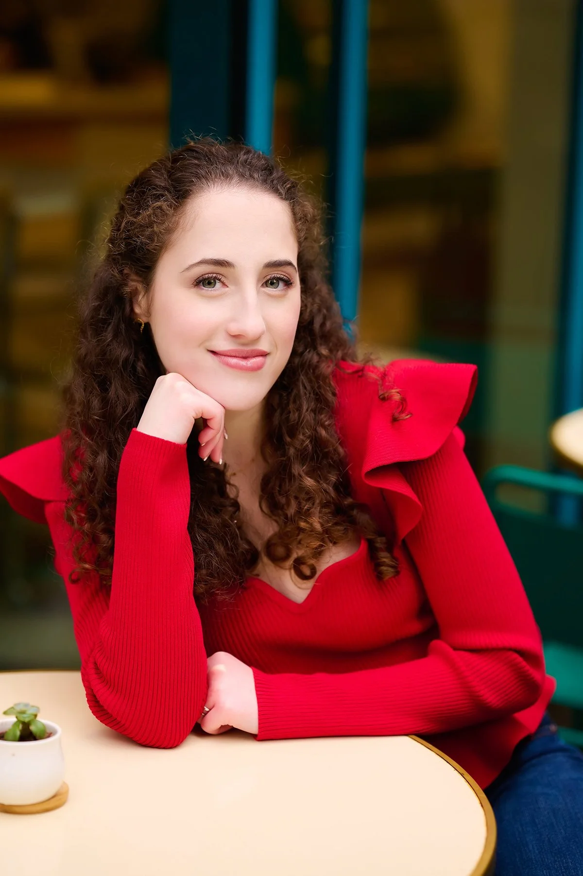 Redhead teen girl long curly hair in ruffled Red top posing seated at cafe.
