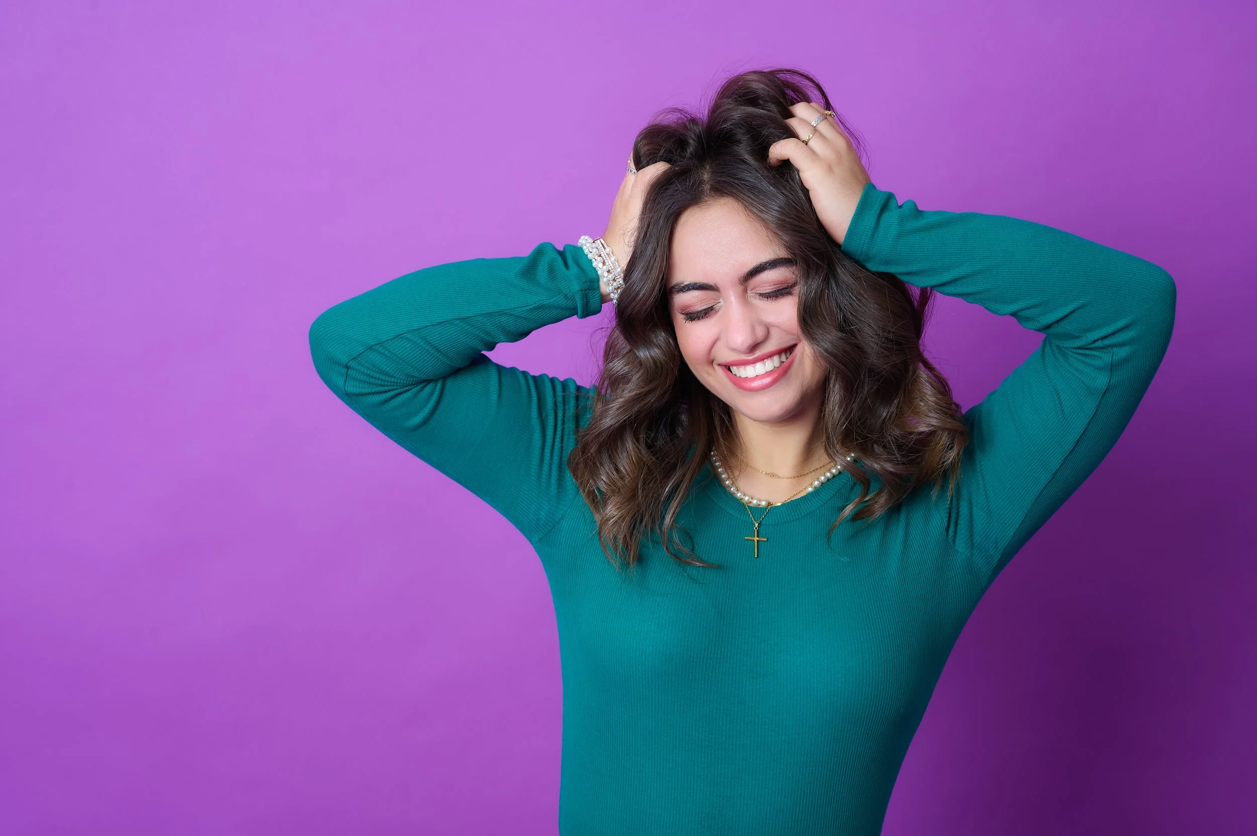 A young woman with brown hair styled in loose waves, smiling and with her eyes closed, wearing a teal long-sleeved shirt, jewelry including bracelets, a necklace with a cross pendant, and a ring, standing against a purple background.