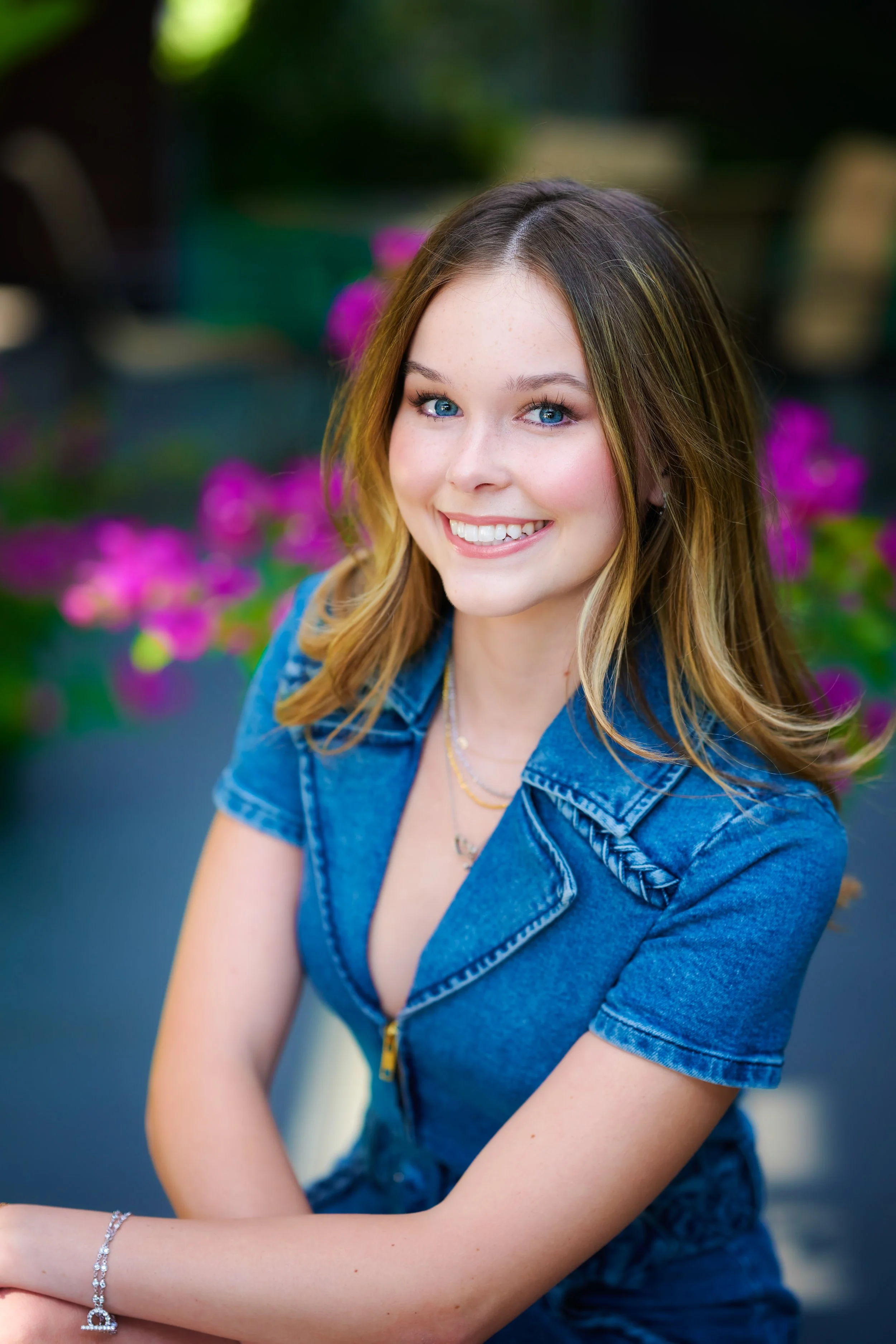 A young woman with blue eyes and light brown, wavy hair smiles while sitting outdoors among pink flowers, wearing a denim zip-up shirt and jewelry.