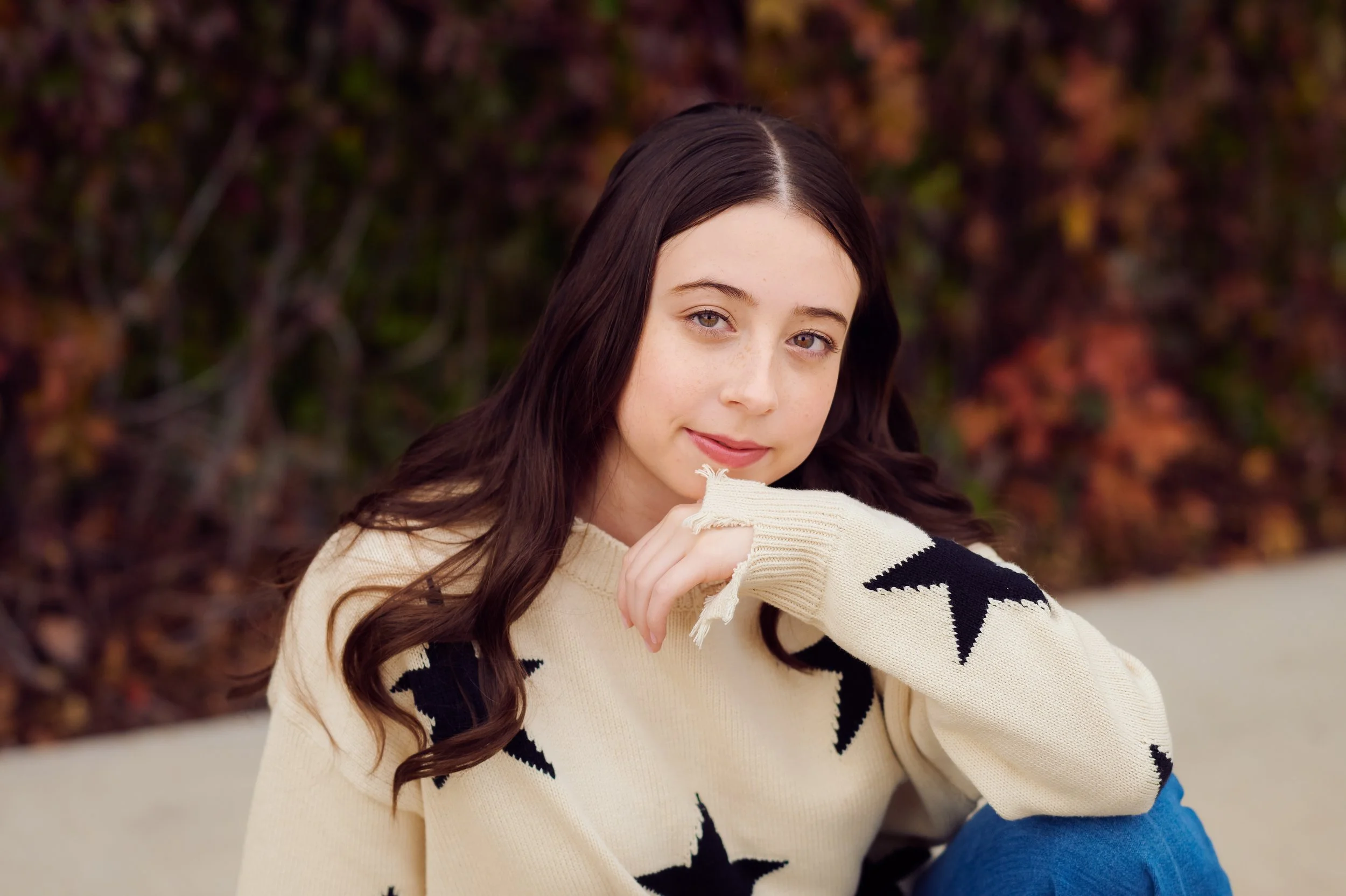 A young woman with long dark hair and fair skin, wearing a cream sweater with black star patterns and ripped jeans, sitting outdoors in front of autumn foliage.