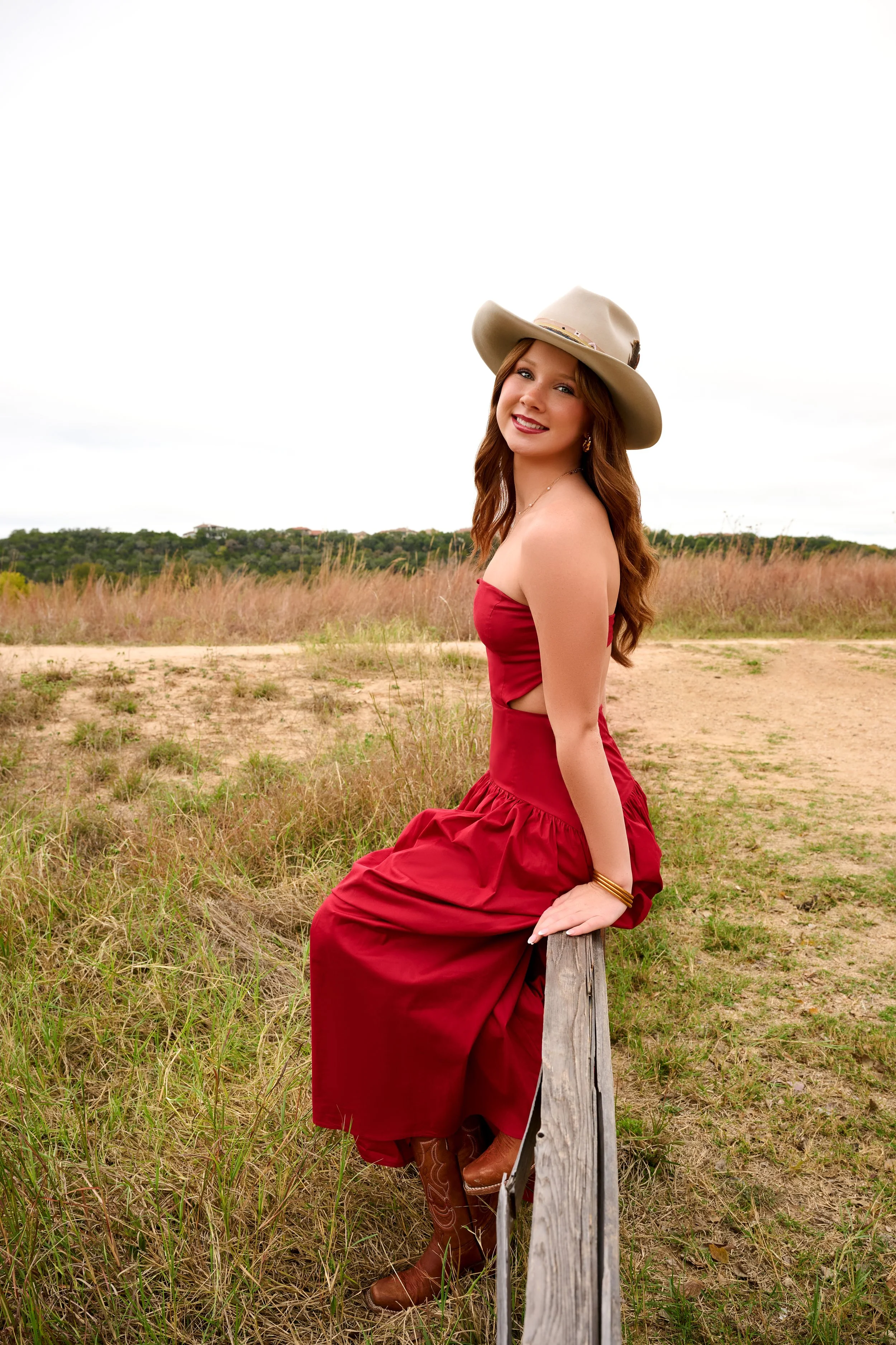 A woman in a red strapless dress and cowboy boots sitting on a wooden fence post in a rural outdoor area, wearing a wide-brimmed hat and smiling at the camera.
