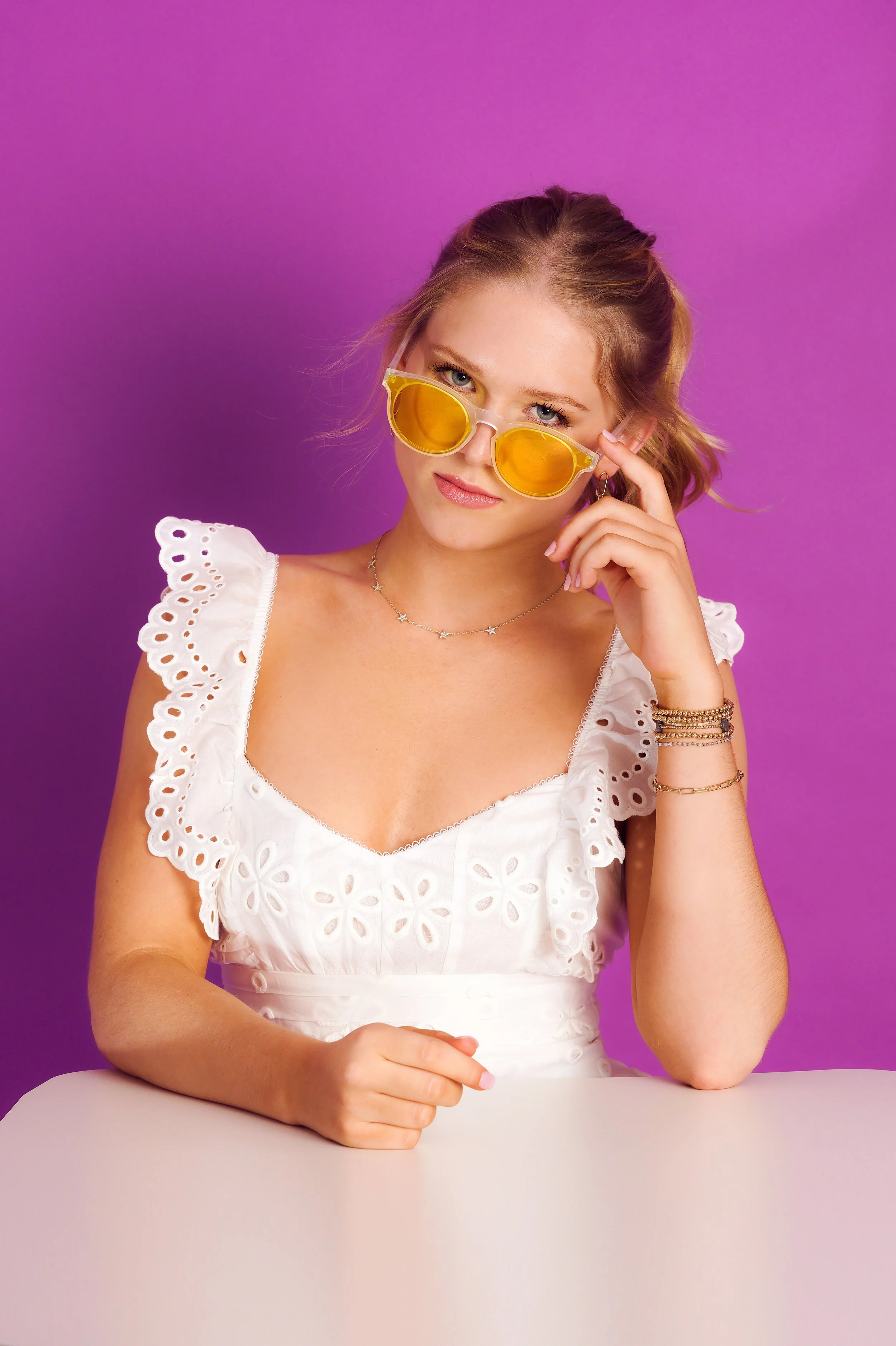 Young woman with blonde hair wearing yellow sunglasses, a white eyelet dress with ruffled sleeves, and layered jewelry, sitting at a table against a purple background.