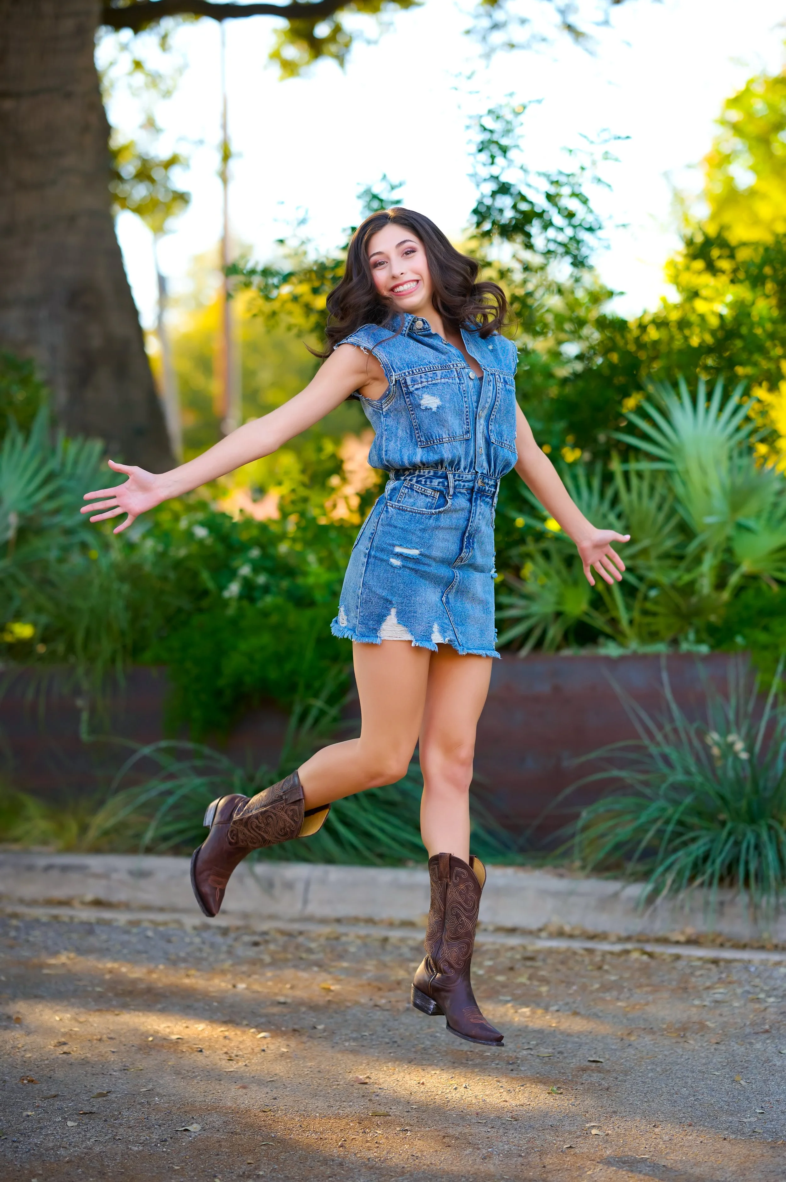 A young woman in a ripped denim sleeveless shirt and skirt jumps joyfully outdoors, wearing cowboy boots with a big smile, surrounded by green plants and trees.
