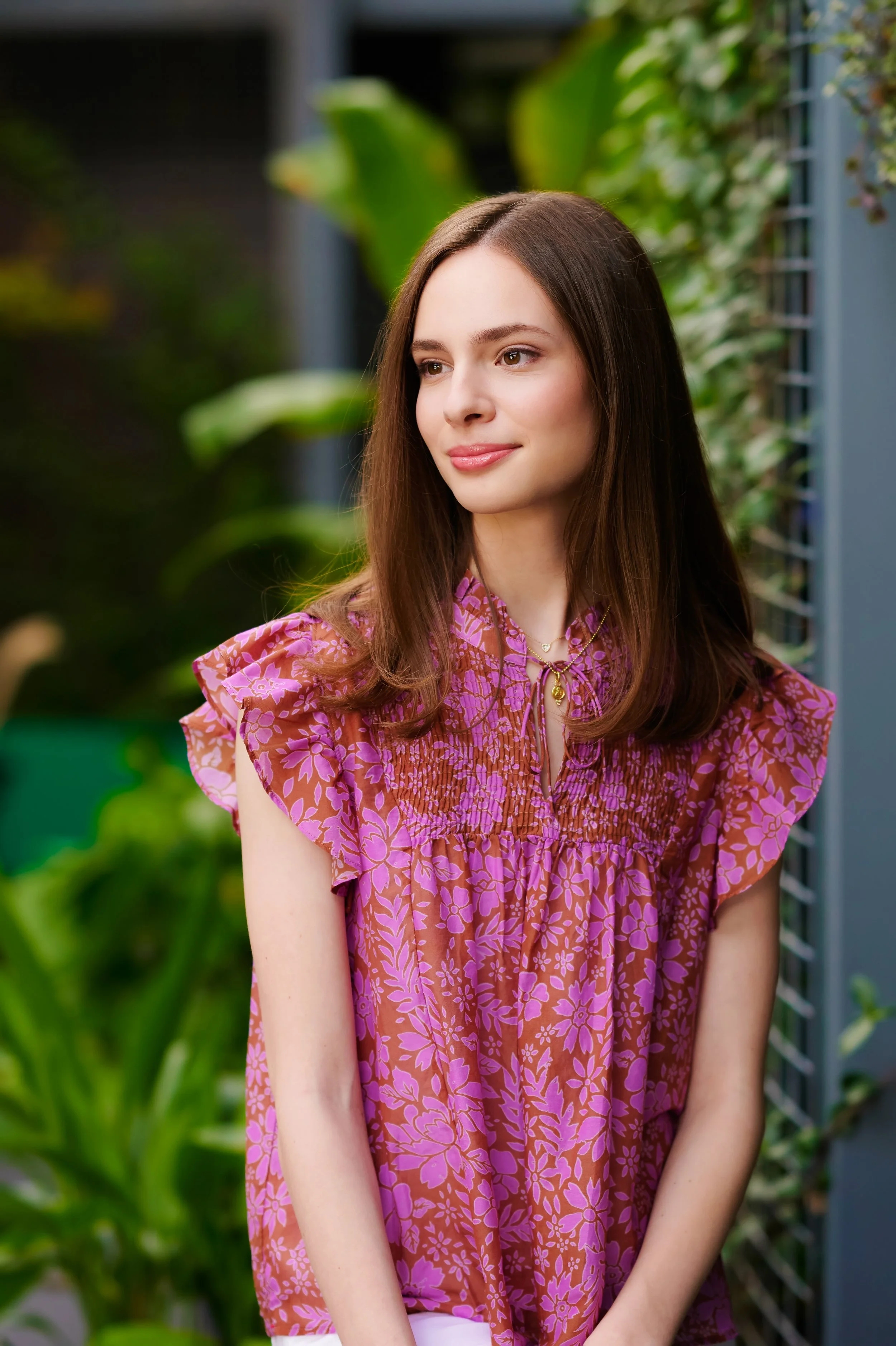 A young woman with long brown hair, wearing a pink floral dress, standing outdoors with lush green plants in the background.