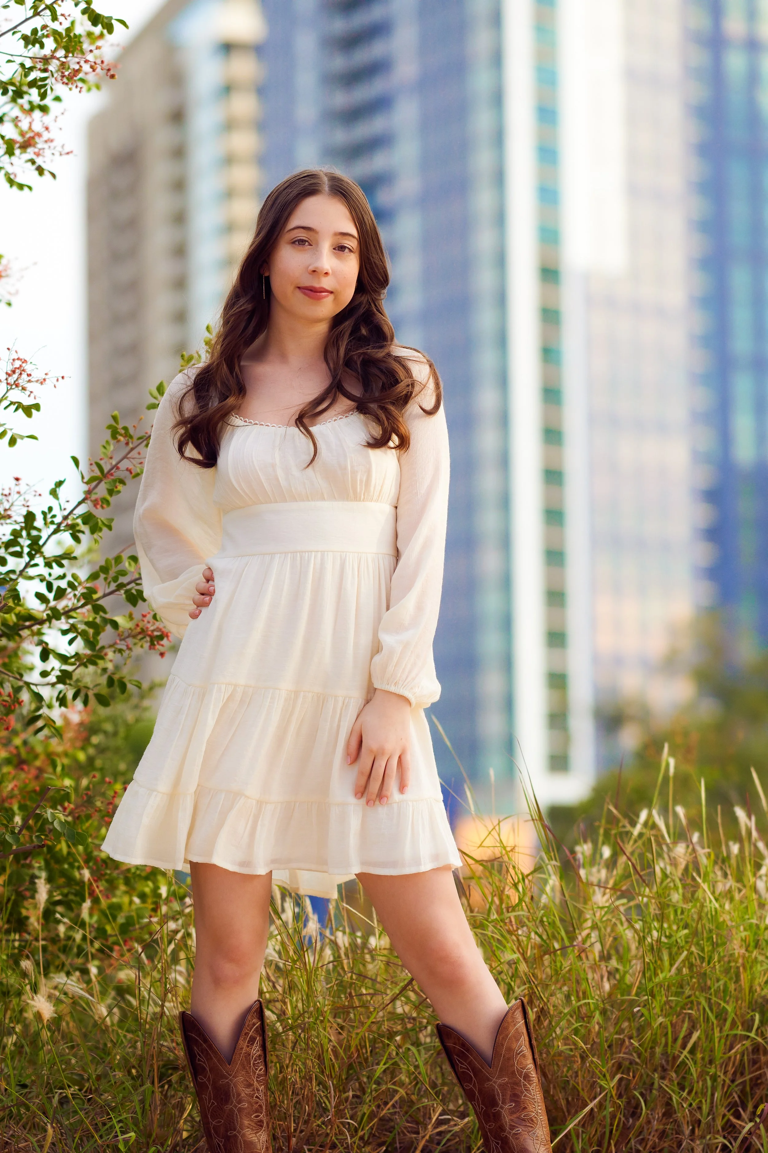 Young woman in a white dress and cowboy boots standing outdoors with tall grass and trees, city buildings in the background, during daytime.