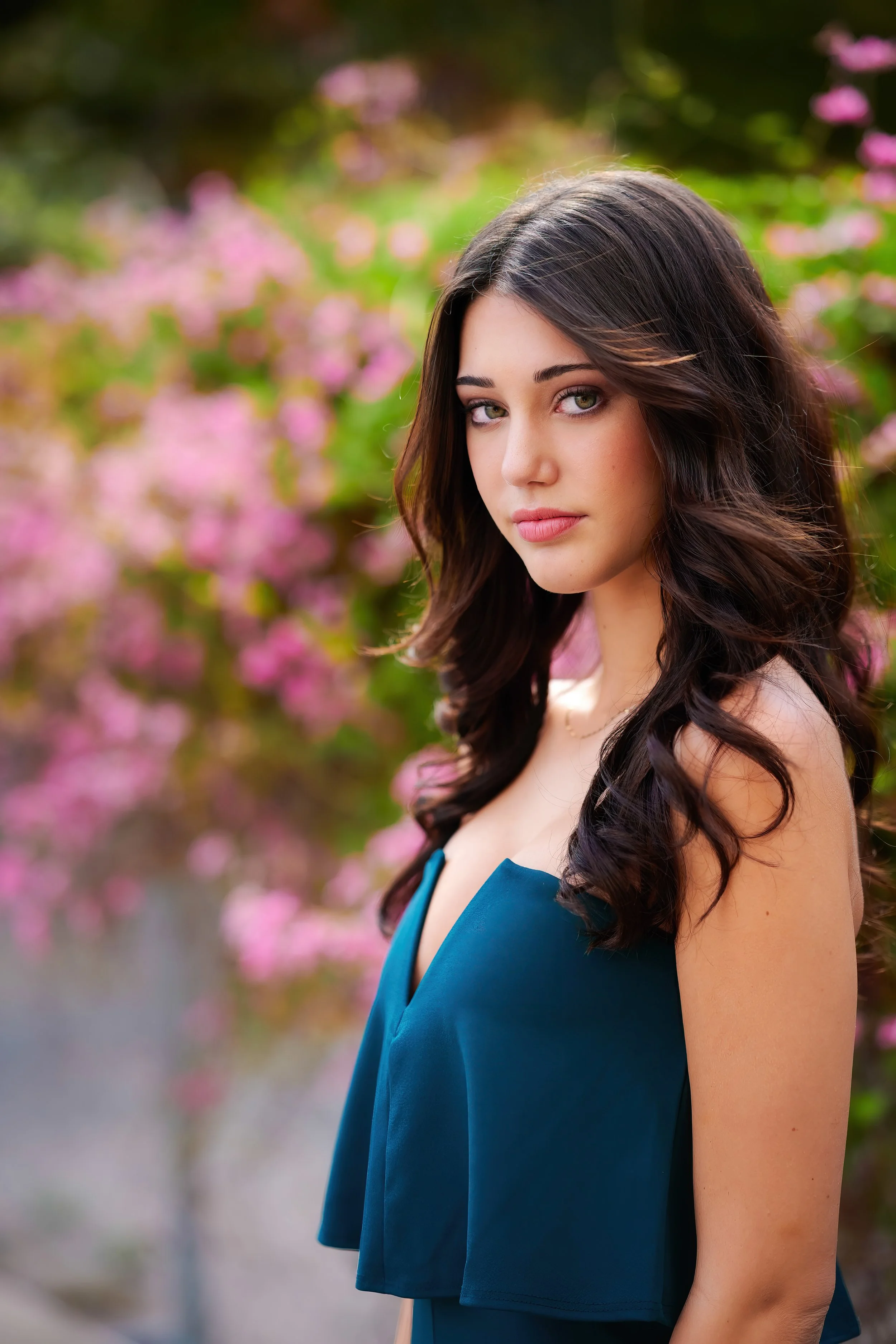 A young woman with long dark hair, wearing a teal dress, standing outdoors with pink flowering bushes in the background.