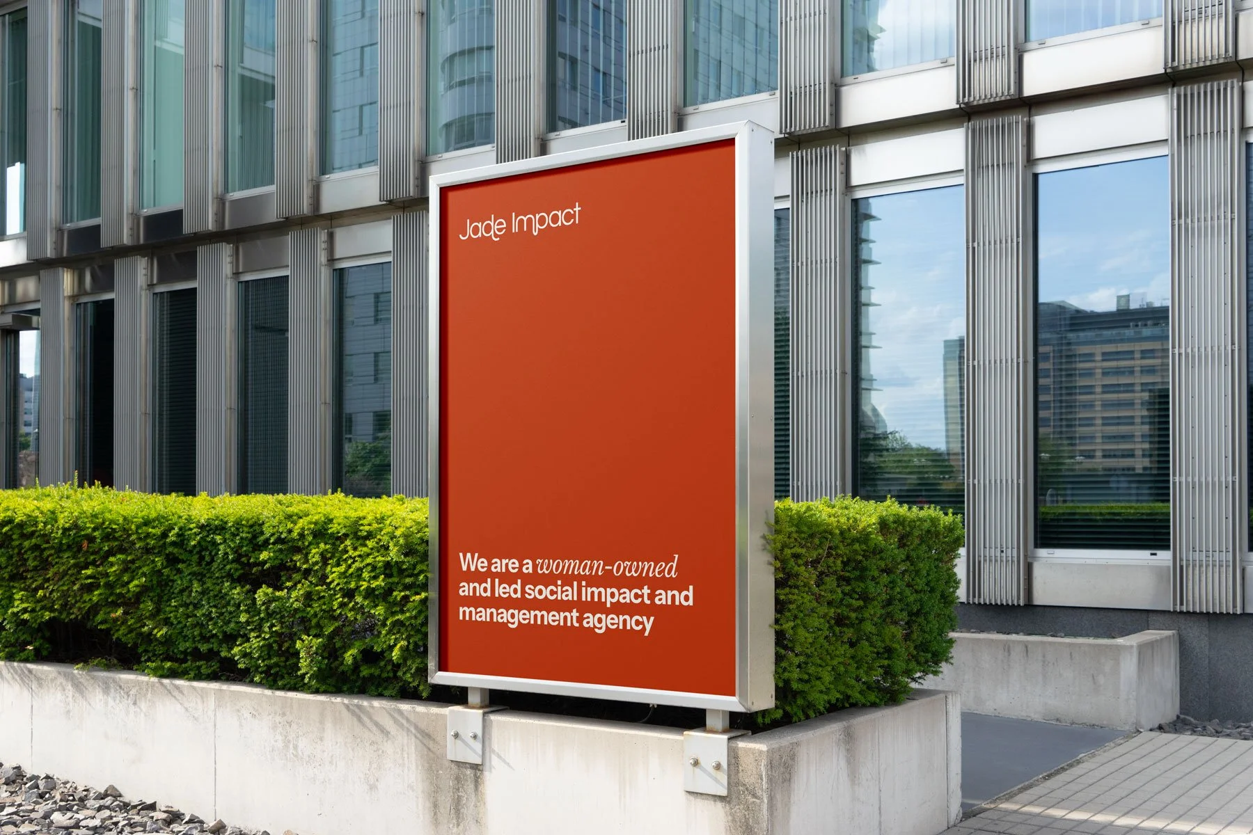 A red signboard outside a modern office building with glass windows and vertical metallic panels. The signboard reads: "Jade Impact" and describes the company as woman-owned, focused on social impact and management.