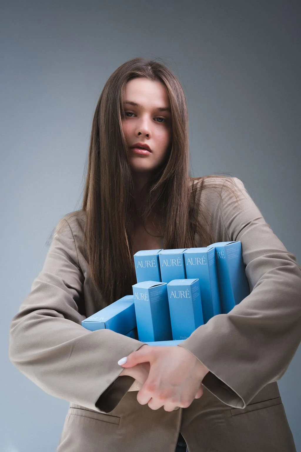 A woman with long brown hair is holding multiple blue boxes of Aure skincare products, wearing a beige blazer, standing against a plain gray background.