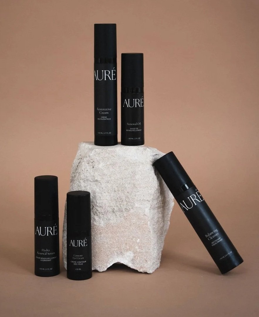Five black skincare product bottles with white text, displayed around a large, white stone against a beige background.