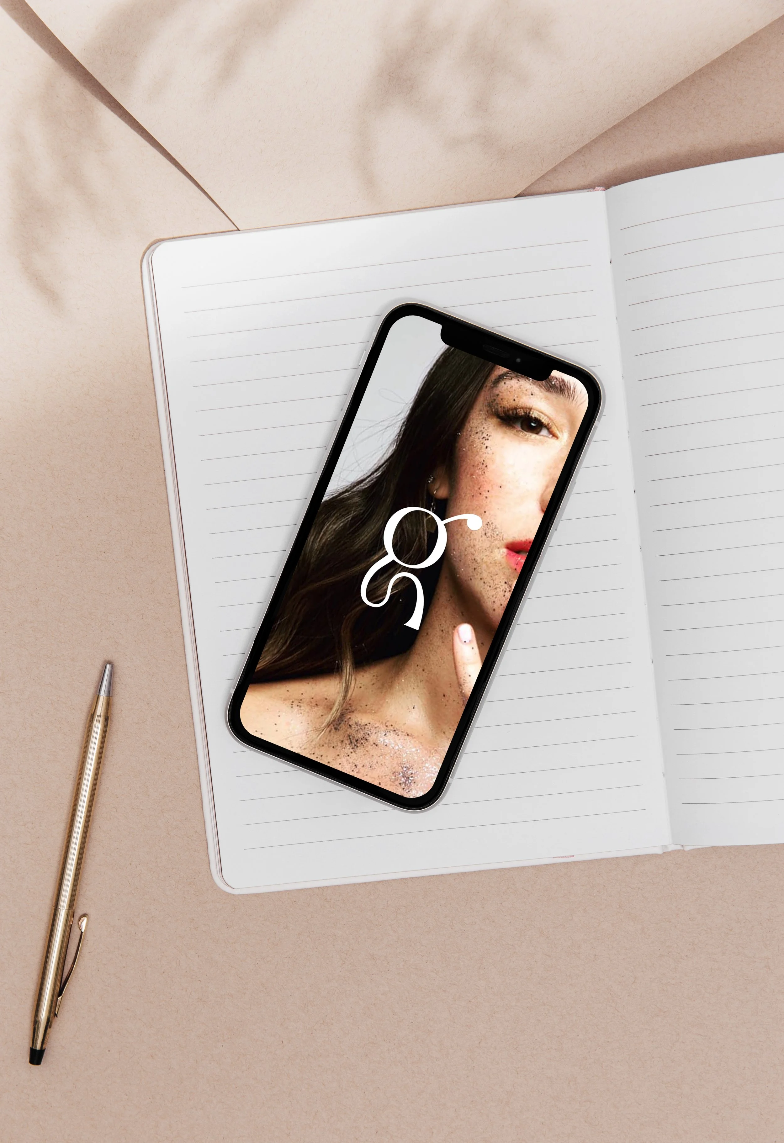 Open notebook with lined pages, a smartphone displaying a woman's face with freckles, and a gold pen on a beige surface.