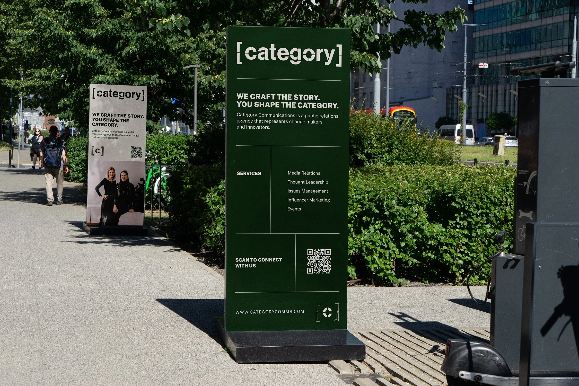 Outdoor sidewalk with green informational sign for Category Communications, listing services like media relations and influencer marketing, with a QR code at the bottom for connection.