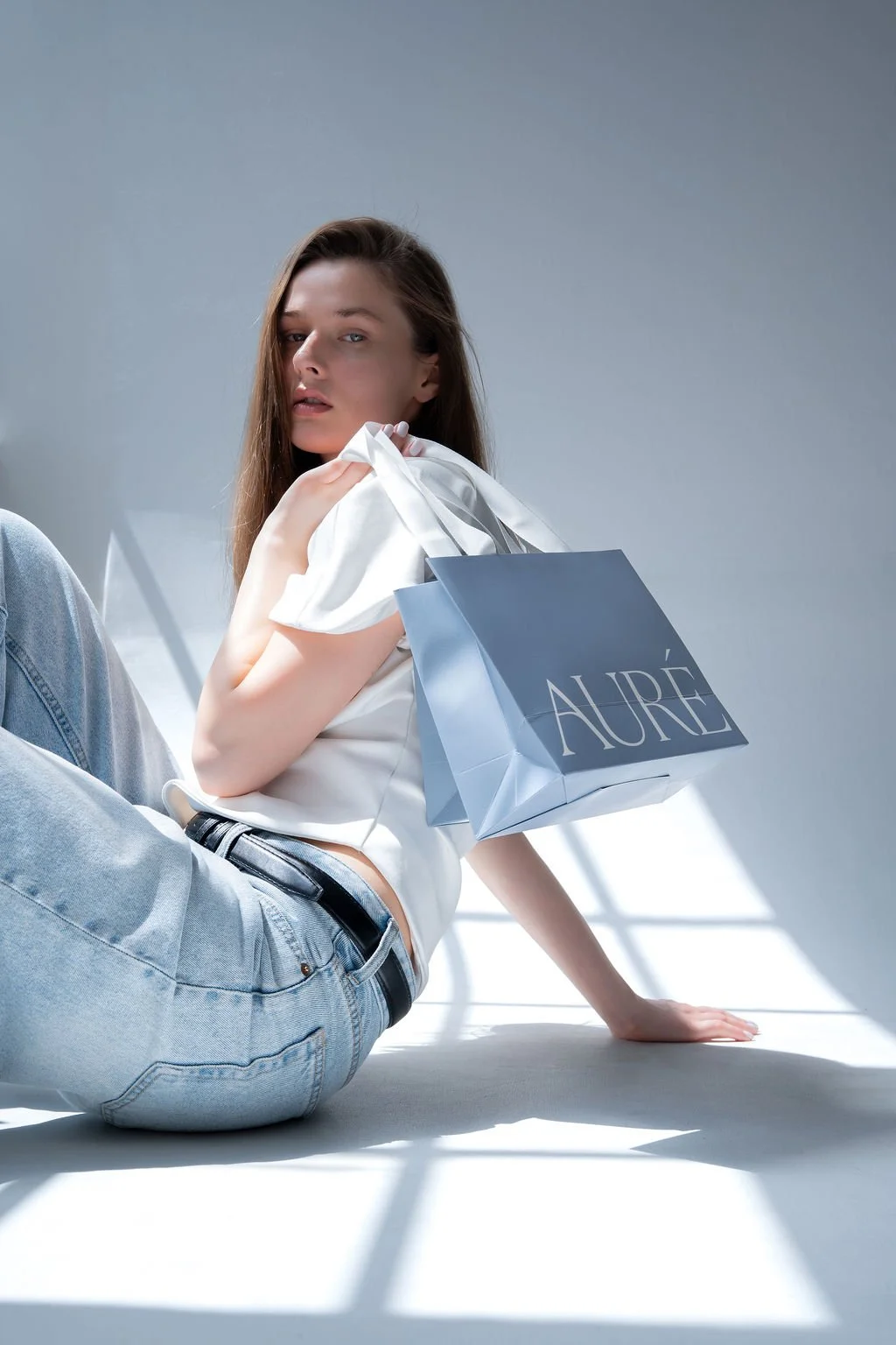 A young woman with brown hair, wearing a white top and light blue jeans, is sitting on the floor holding a shopping bag labeled 'AURE'.