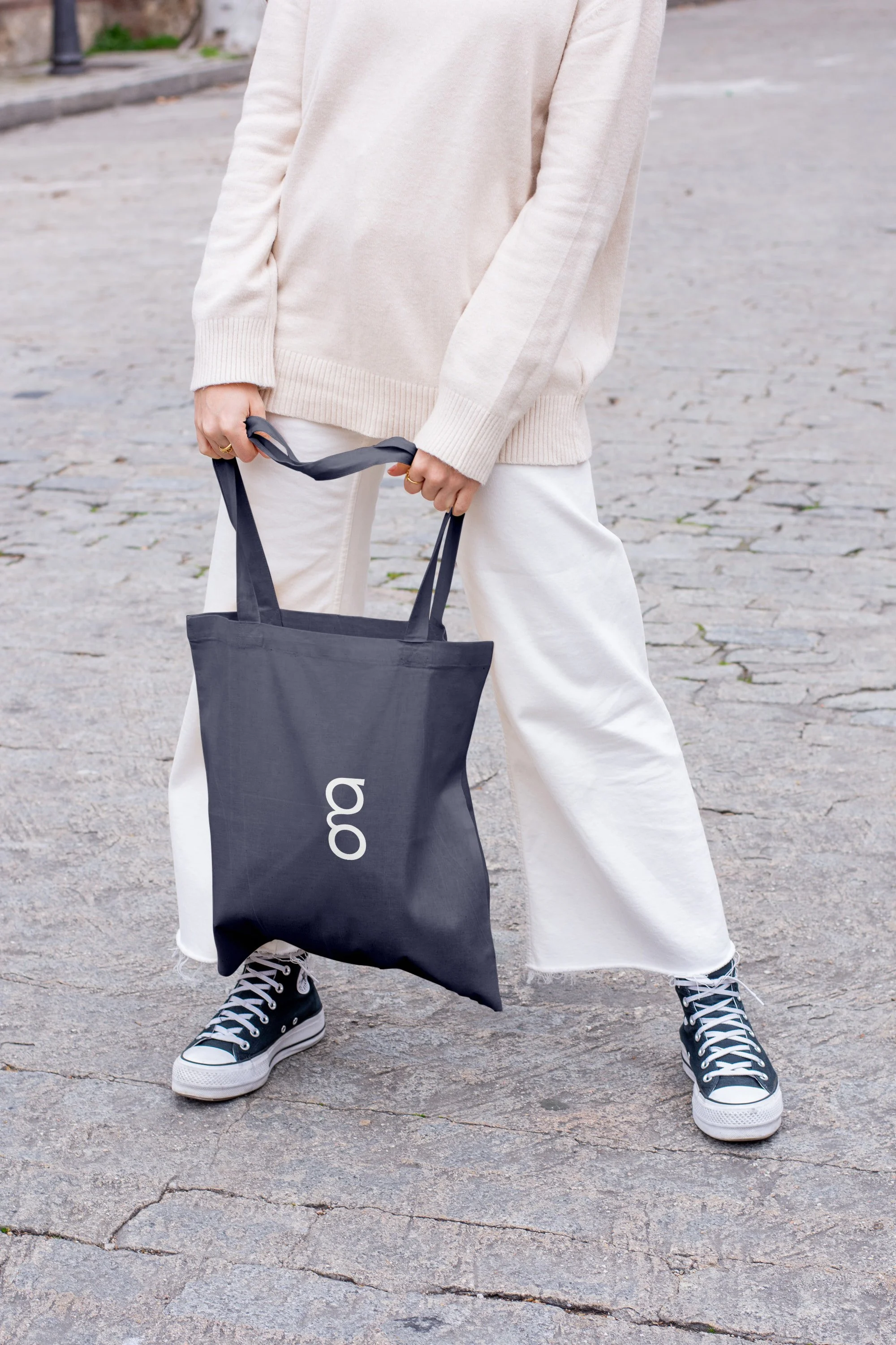 Person holding a black tote bag with white text, wearing black and white sneakers, cream-colored pants, and a beige sweater, standing on a cobblestone street.