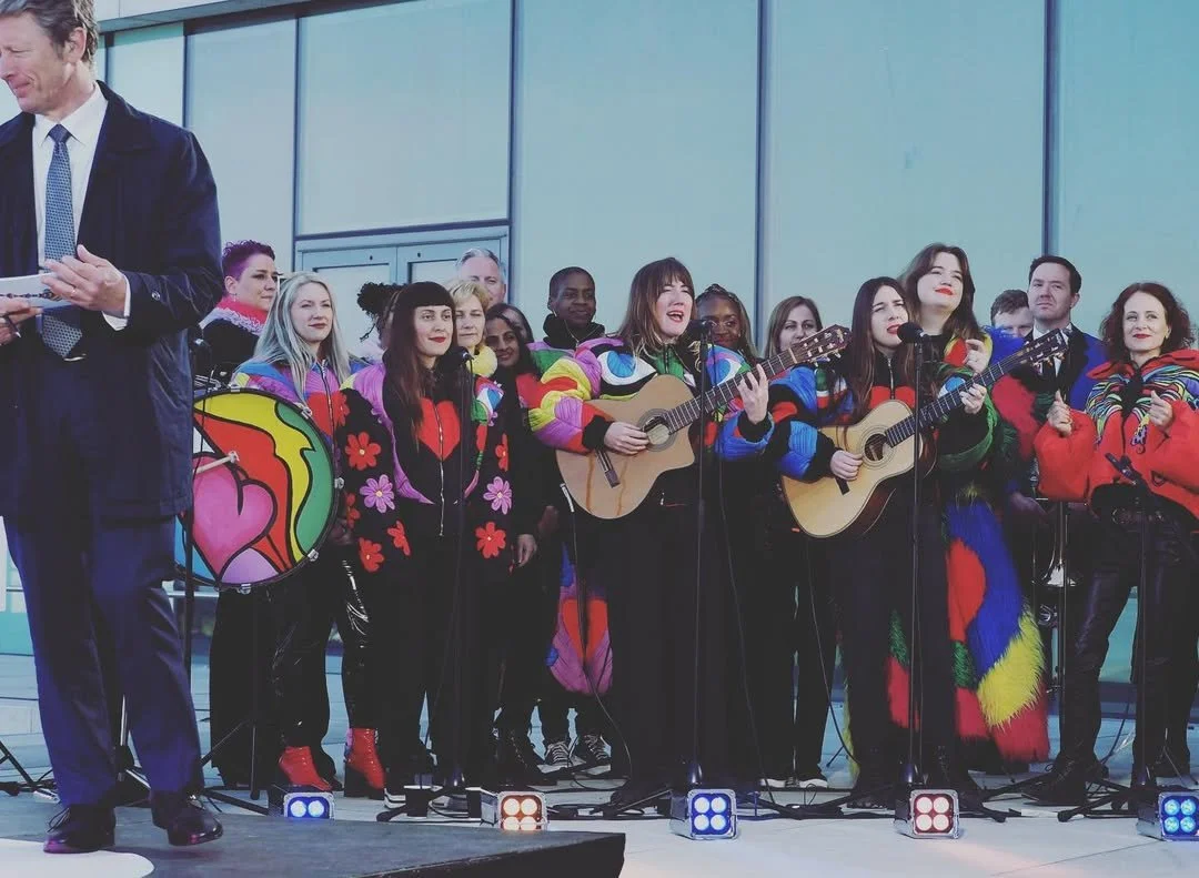 Group of diverse people performing music with guitars and drums, wearing colorful, patterned sweaters, in an indoor setting.