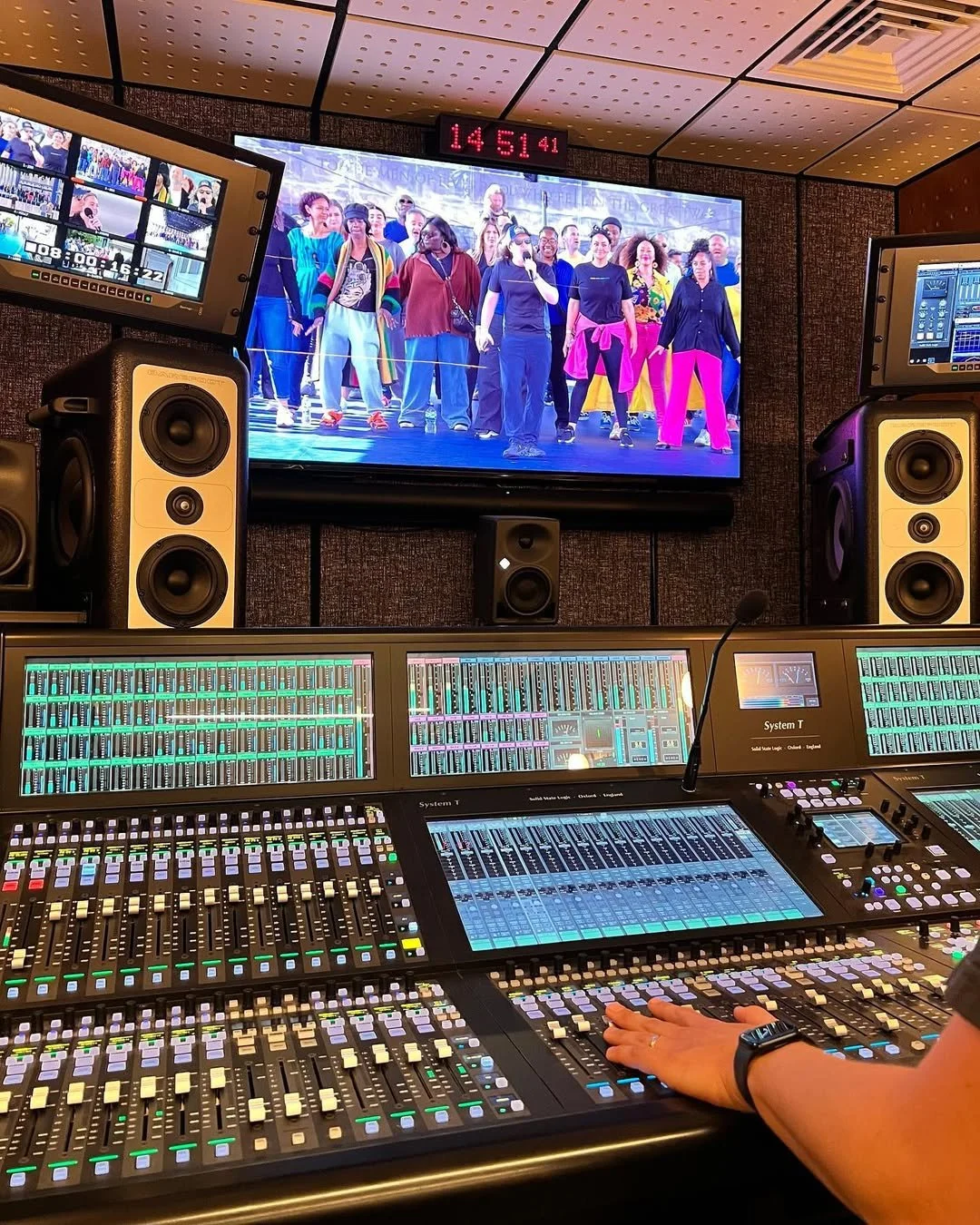 Recording studio with a soundboard and two large speakers in front of a wall-mounted display showing a live rehearsal or performance of a diverse group of people on stage. The display shows a countdown timer, and additional monitors and equipment are visible within the studio.