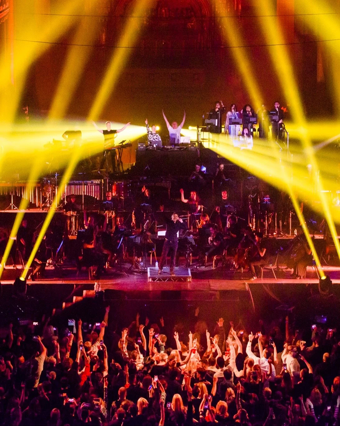 A live concert with musicians and singers performing on stage. The audience is cheering with hands raised, and bright yellow stage lights illuminate the scene.