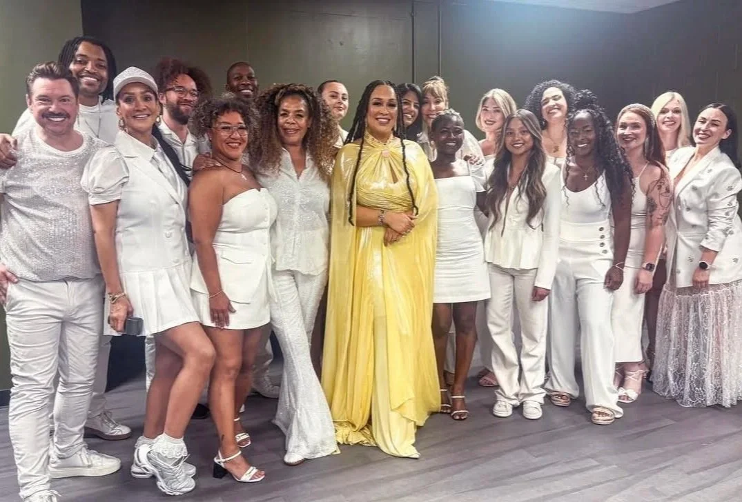 Group of diverse women and men celebrating together indoors, dressed in mostly white and one woman in a yellow dress.