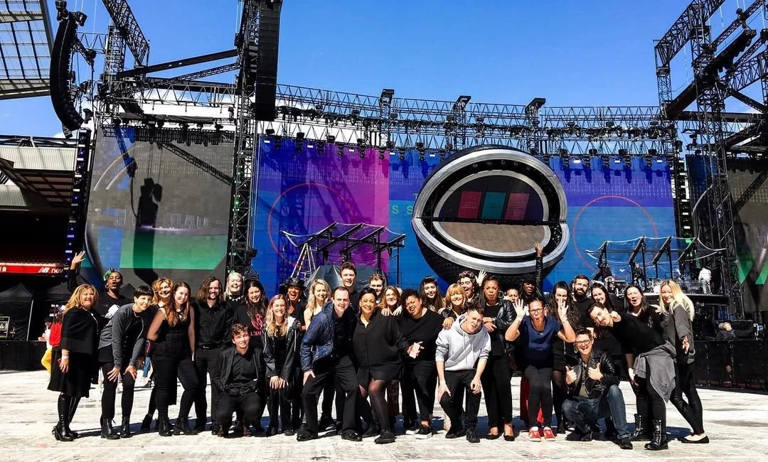 A group of people posing on a stage in front of a large outdoor stage setup during daytime.