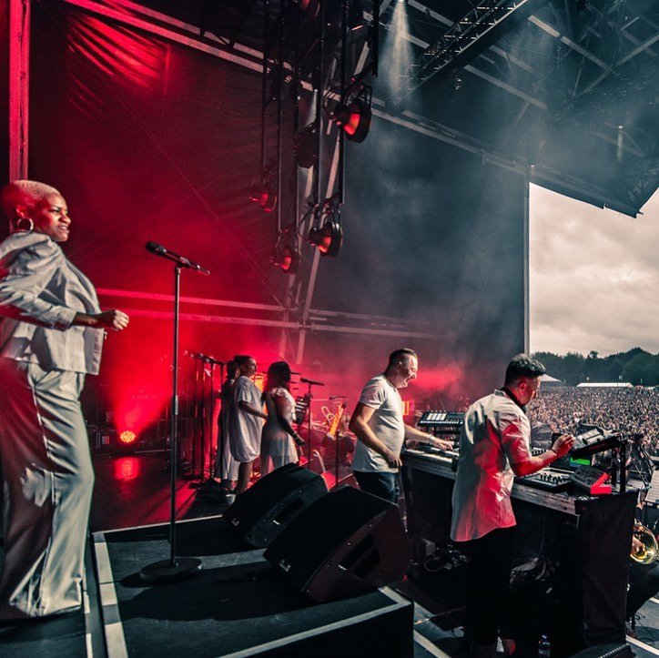 Performers and musicians on stage at a large outdoor concert with red lighting and a crowd in the background.