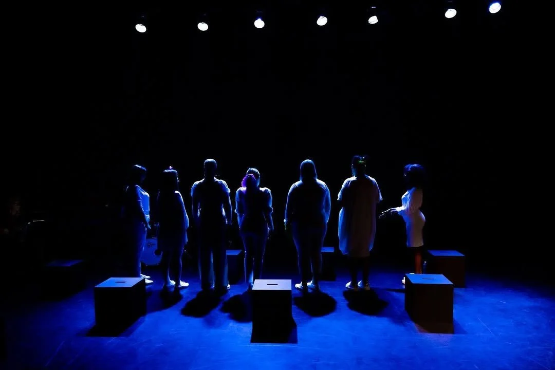 A group of people standing on a stage with their silhouettes illuminated by overhead stage lights.