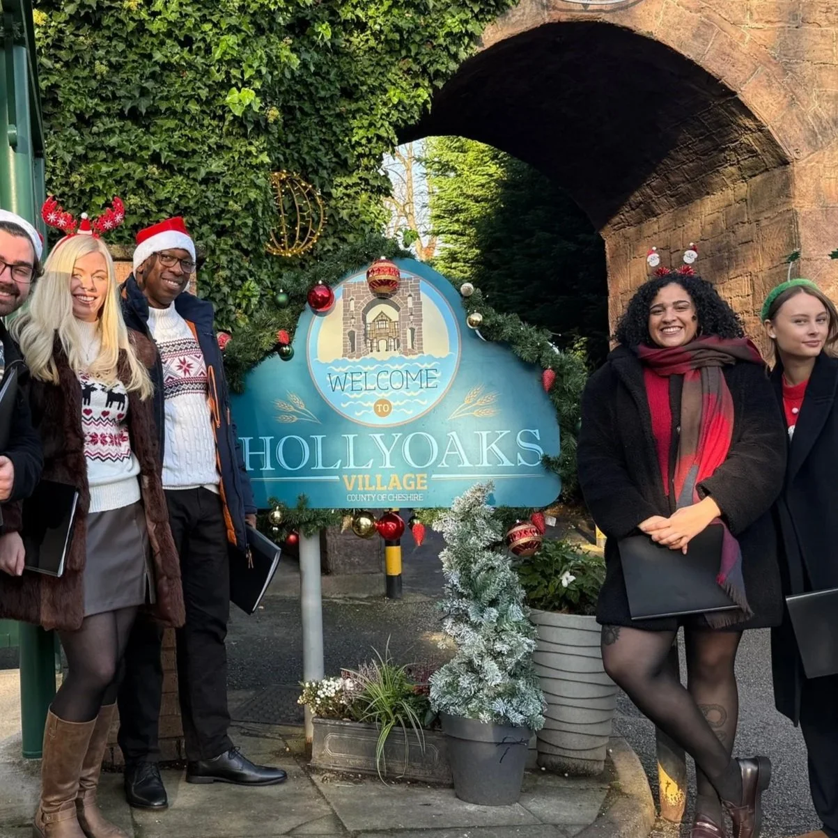 Six people dressed in festive holiday attire standing around a sign that says 'Welcome to Hollyoaks Village, County of Cheshire' decorated with Christmas ornaments and greenery, under a stone archway with foliage and Christmas decorations.