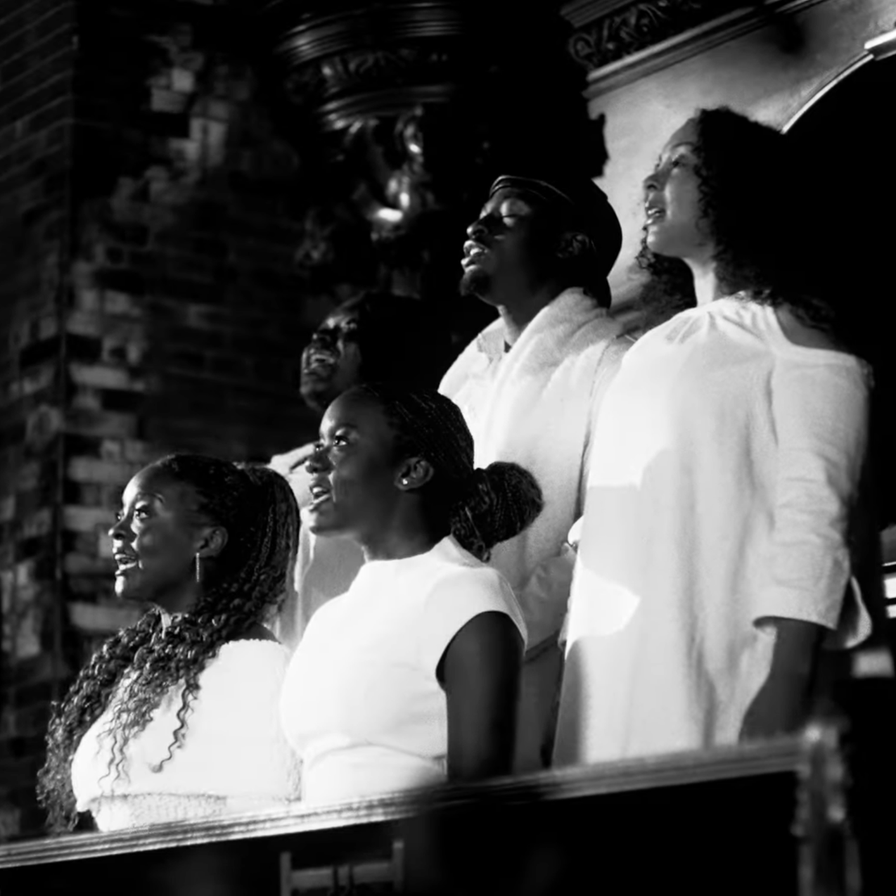 A black-and-white photo of a diverse group of six people sitting in a church or chapel, looking upward with expressions of reverence or prayer.