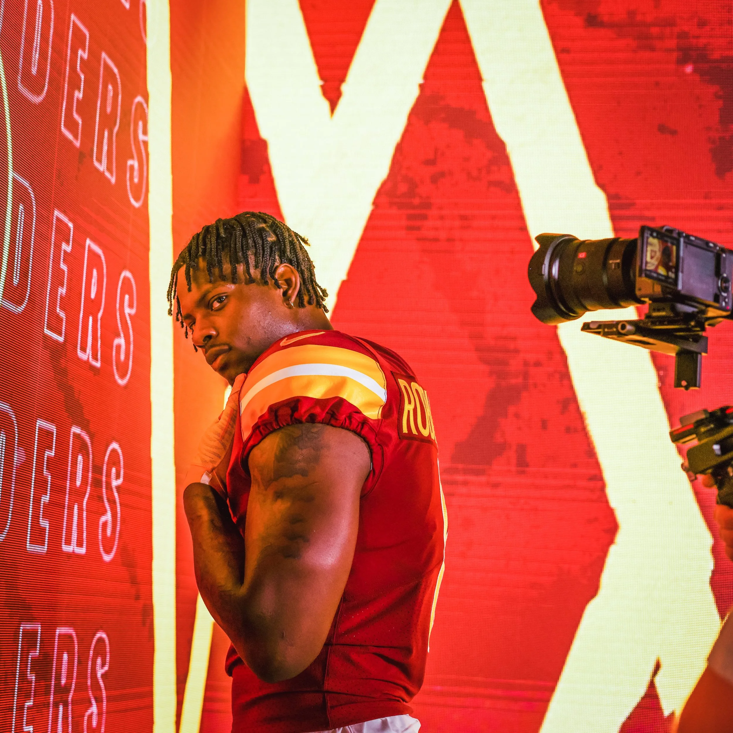 An athlete in a red and yellow football uniform standing in front of a large digital screen with red and yellow graphics, looking over his shoulder.