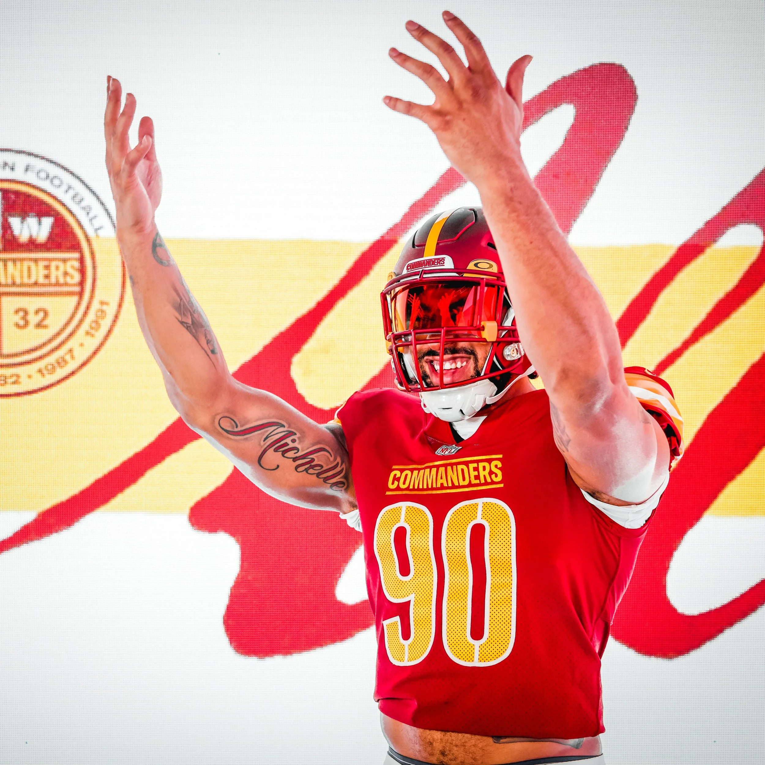 A football player wearing a red and yellow uniform with the number 90 and the word 'Commanders' on it, celebrating with arms raised in front of a backdrop with the Washington Commanders logo.