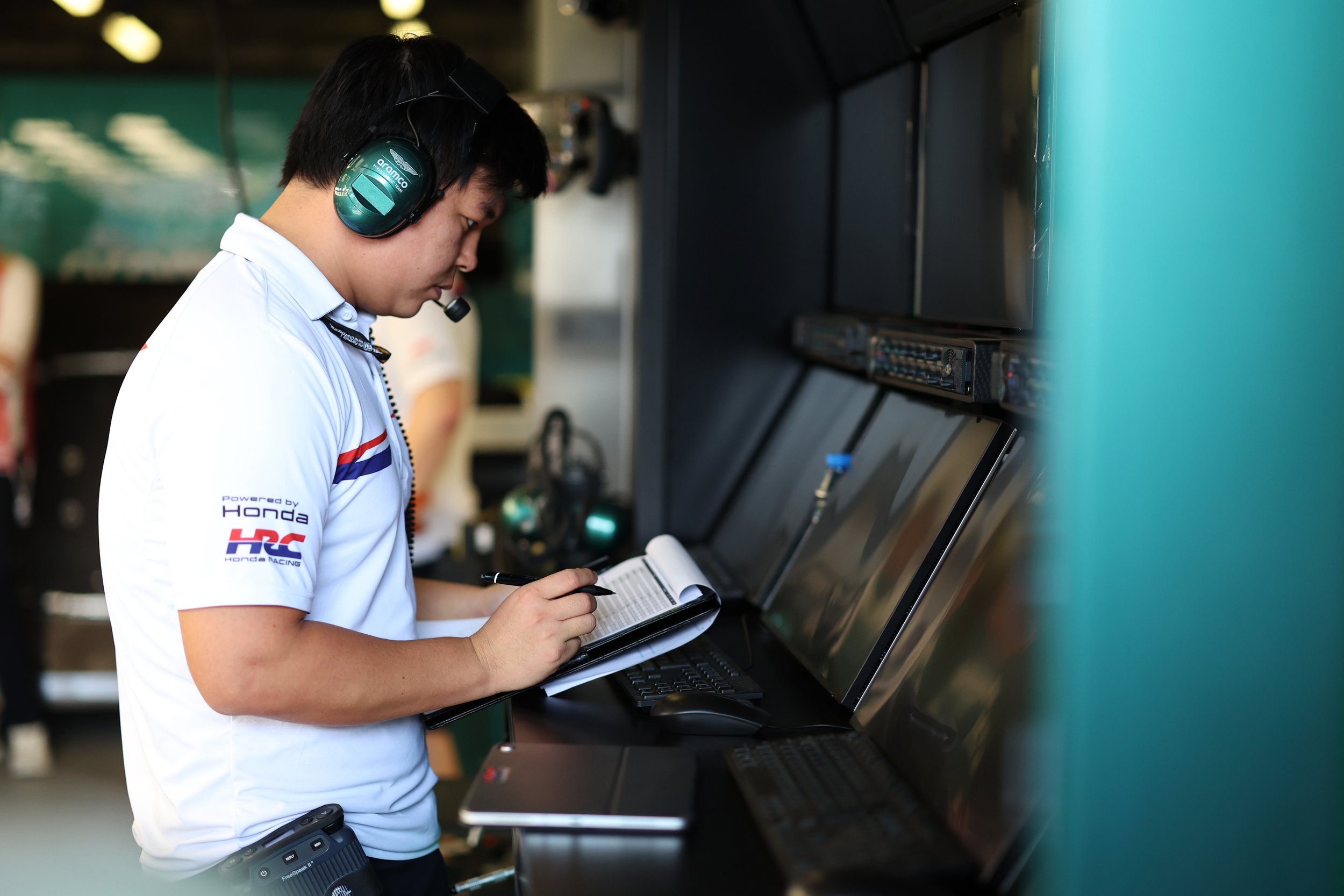 A young man with black hair, wearing a white racing shirt with Honda and racing sponsor logos, is working on a computer at a racing paddock. He is wearing green headphones and appears to be writing in a small notebook.