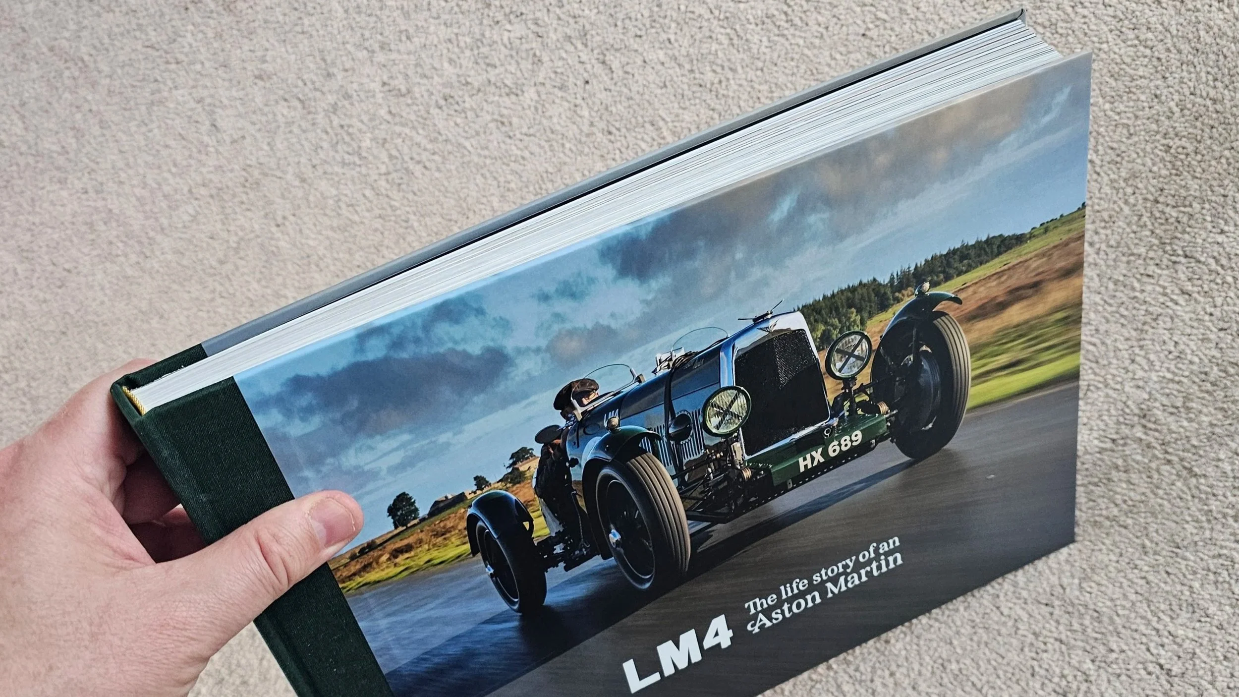 A hardcover book titled 'LM4: The life story of an Aston Martin' with a cover photo of a vintage racing car on a road, under a cloudy sky, being held against a beige carpet.