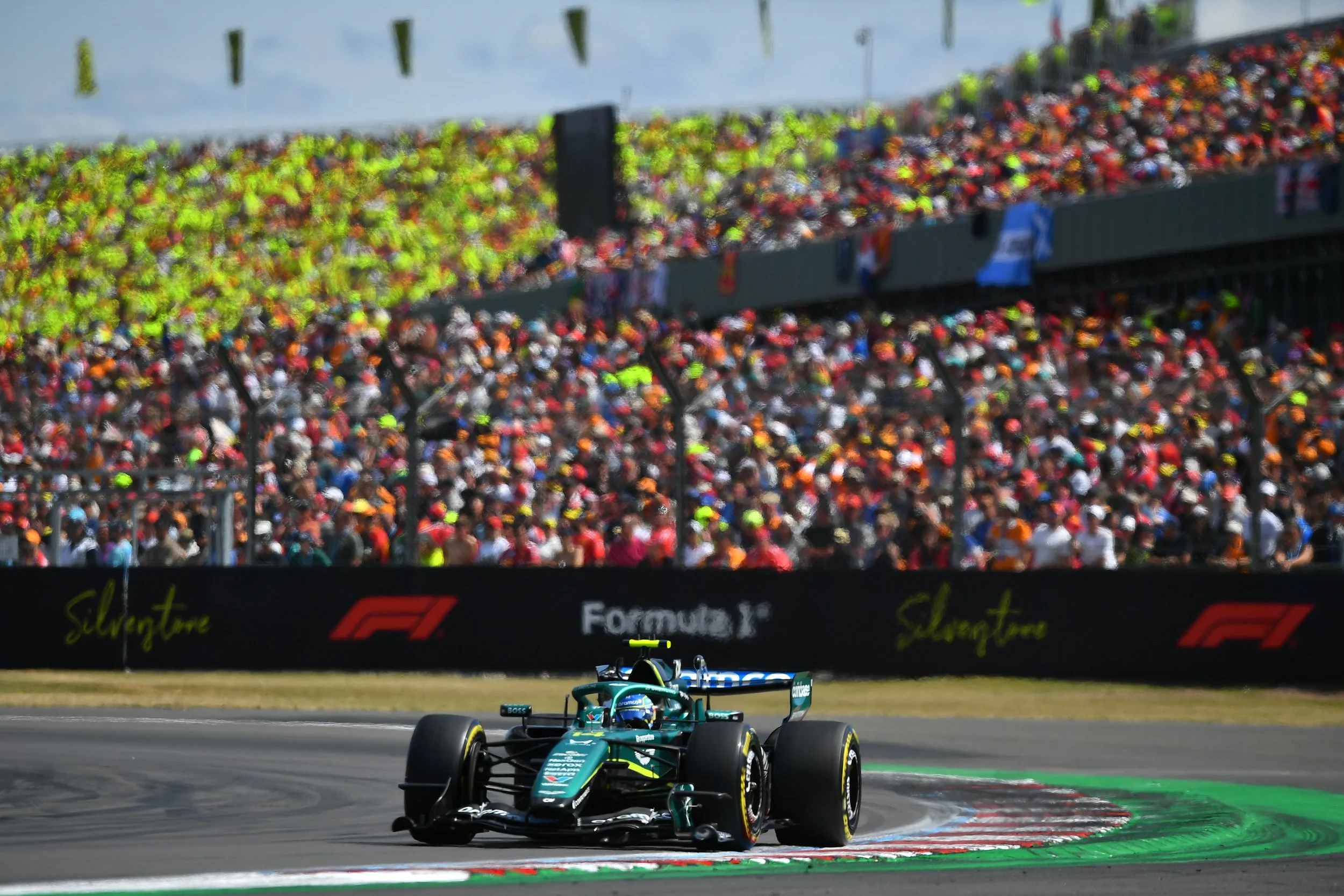 A Formula 1 race car on a race track, approaching a corner with a green and red curb, with a large crowd of spectators in the stands and a barrier with sponsor logos in the background.