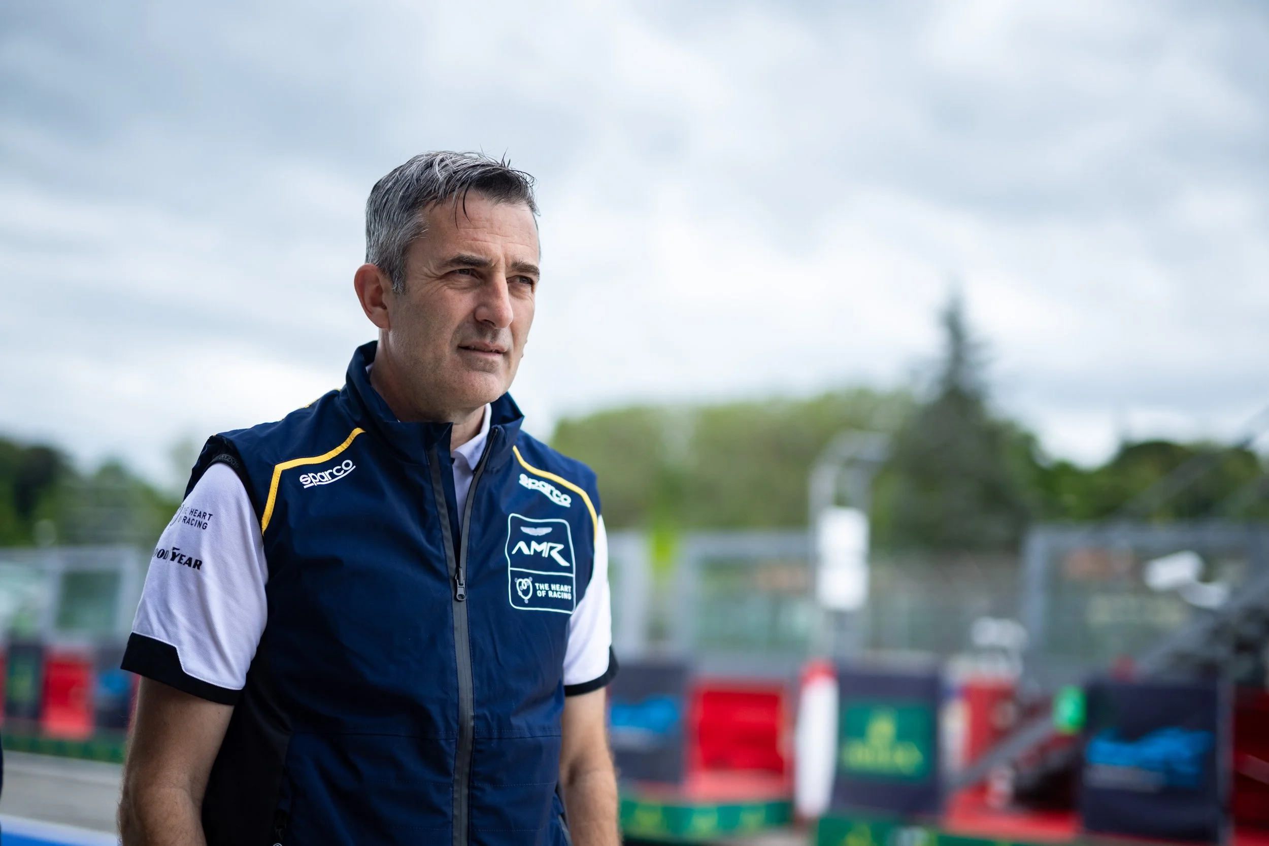 A man in a racing uniform standing outdoors with a race track and cloudy sky in the background.