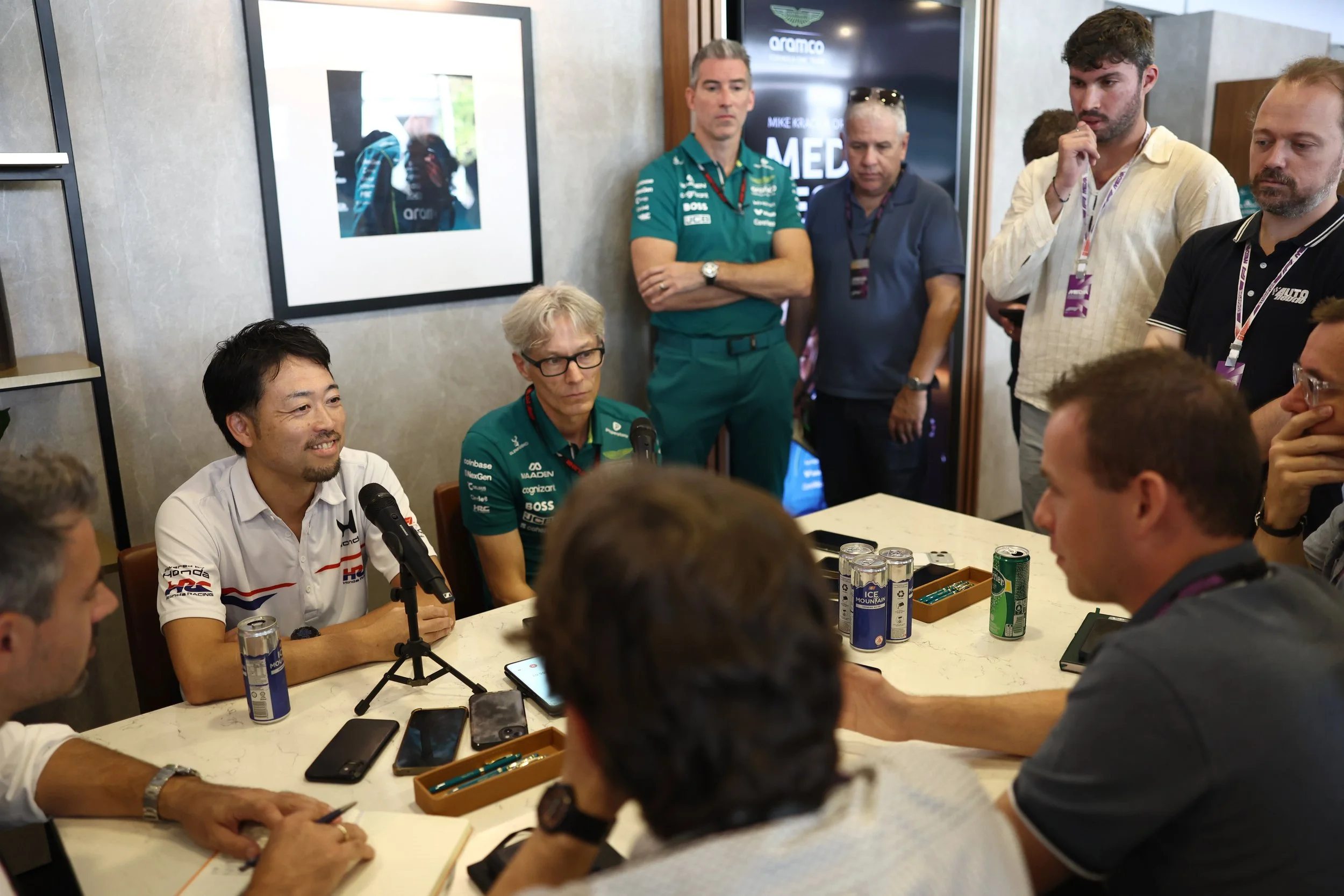 Group of men around a conference table, some with racing team shirts, engaging in a discussion or interview during a media event.