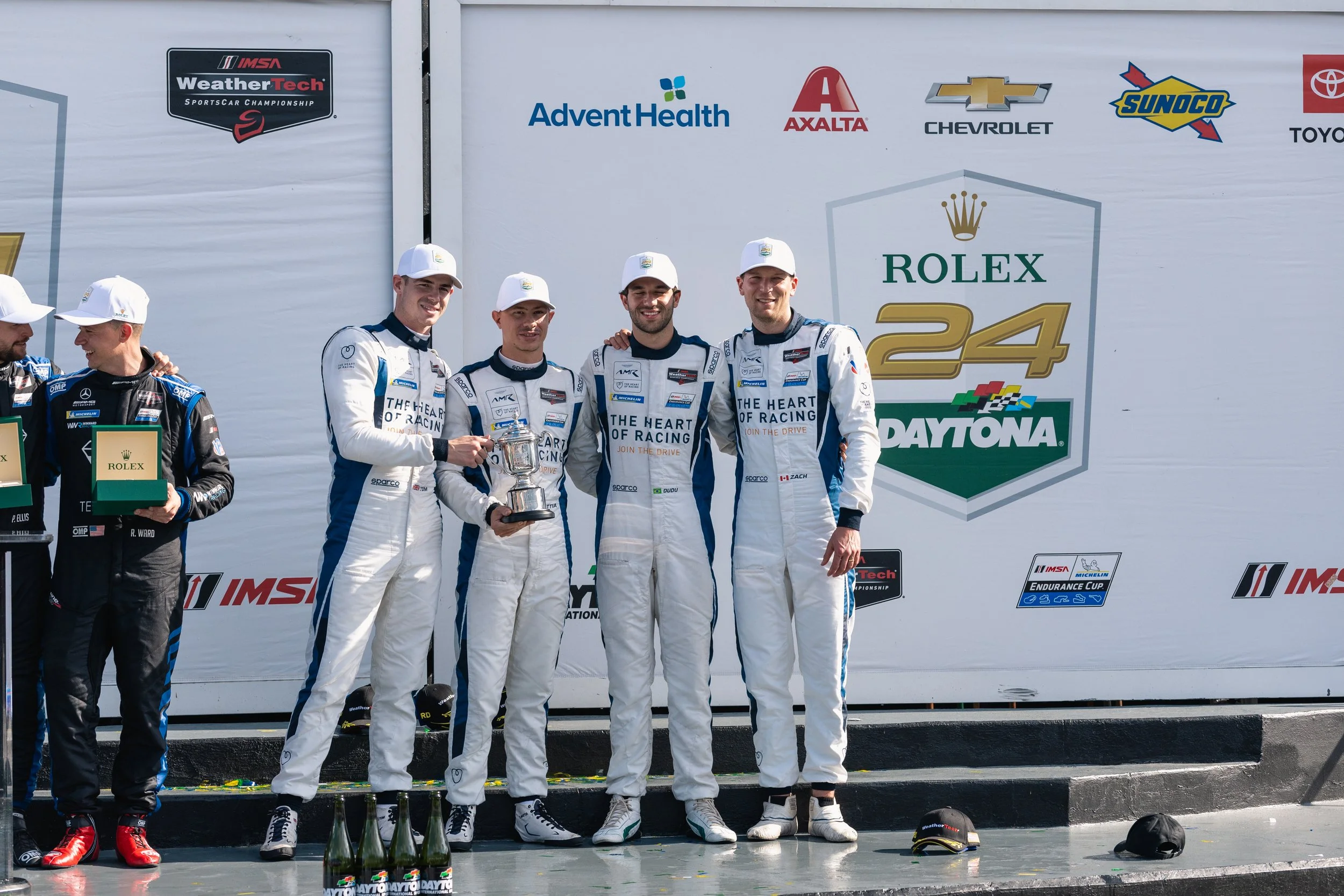 Four race car drivers in white racing suits and white caps standing together on the podium after a race, with trophies and champagne bottles in front of them and sponsor logos on the backdrop behind them.