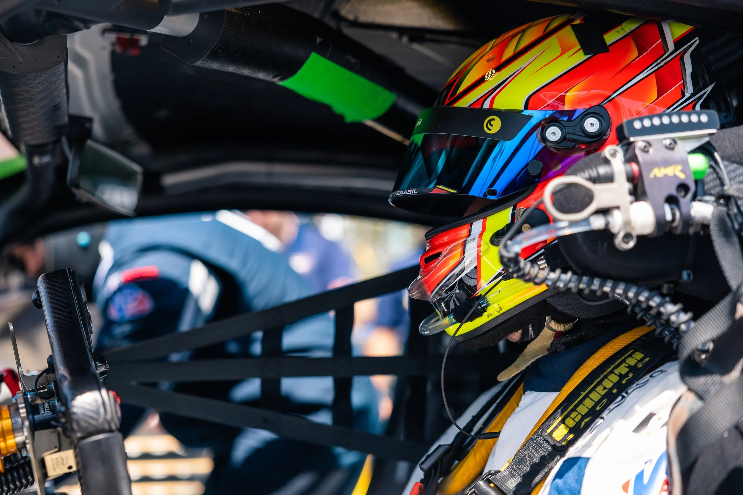 Close-up of a race car driver in a multicolored helmet and racing gear inside an Aston Martin race car, with a blurred background.