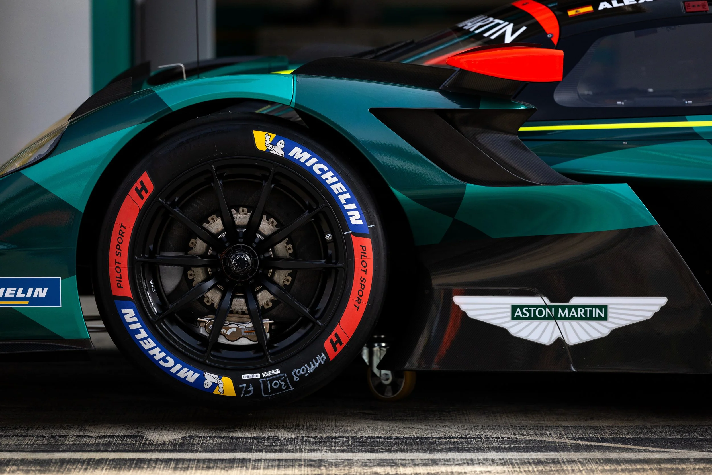 Close-up of a race car's front wheel with Michelin racing tires and a teal and black body, featuring sponsorship decals from Michelin and Aston Martin.