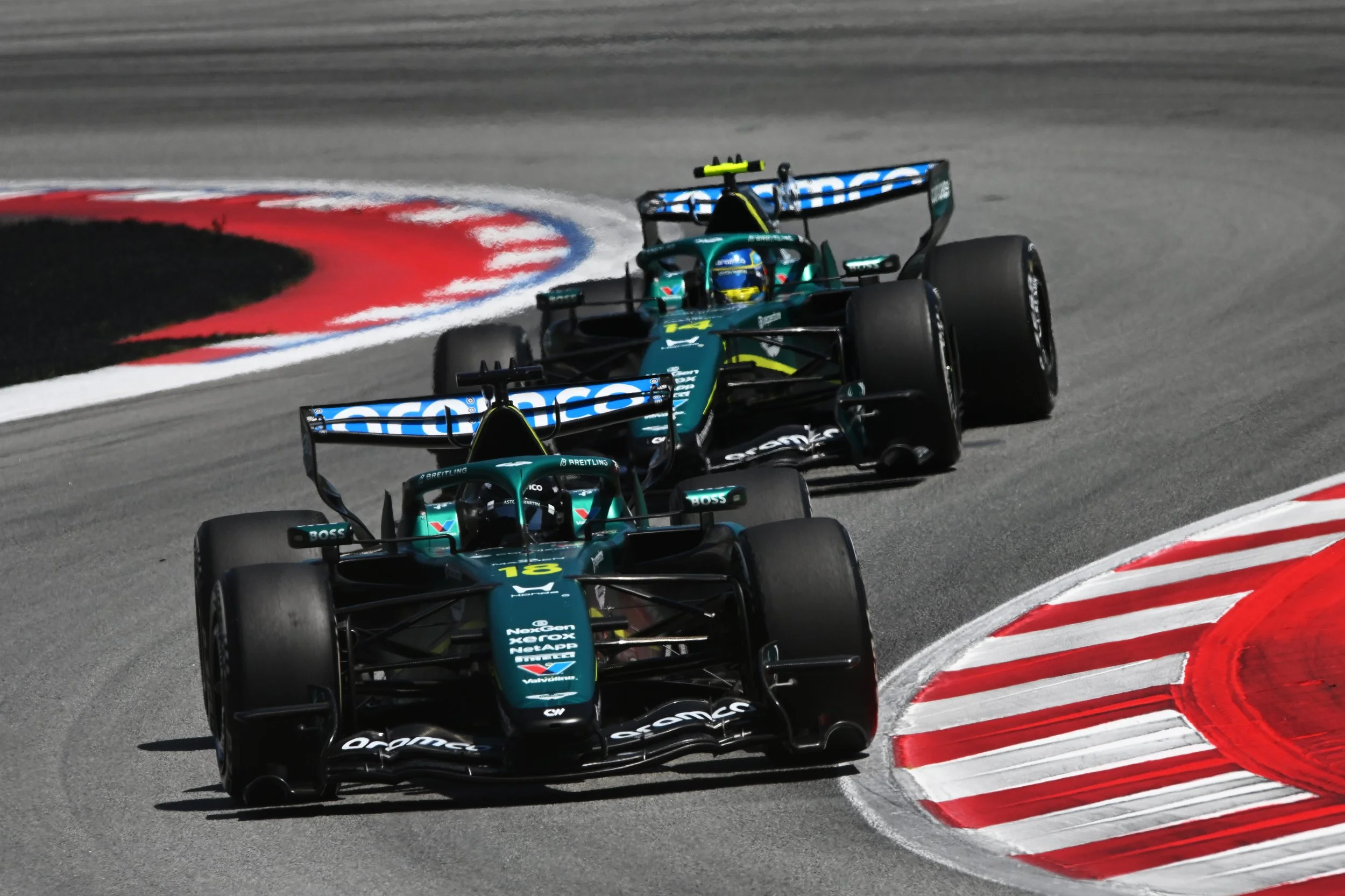 Two Aston Martin AMR26 Formula 1 race cars, being driven by Stroll and Alonso, at the Barcelona Race Track 2026, going around a corner with red and white curbs.