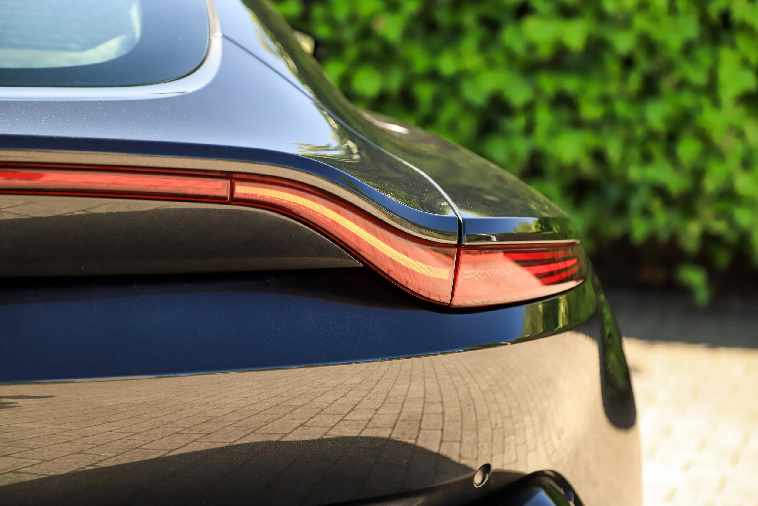 Close-up of the rear end of a sleek black luxury car showing the taillight and a small part of the rear window with greenery in the background.