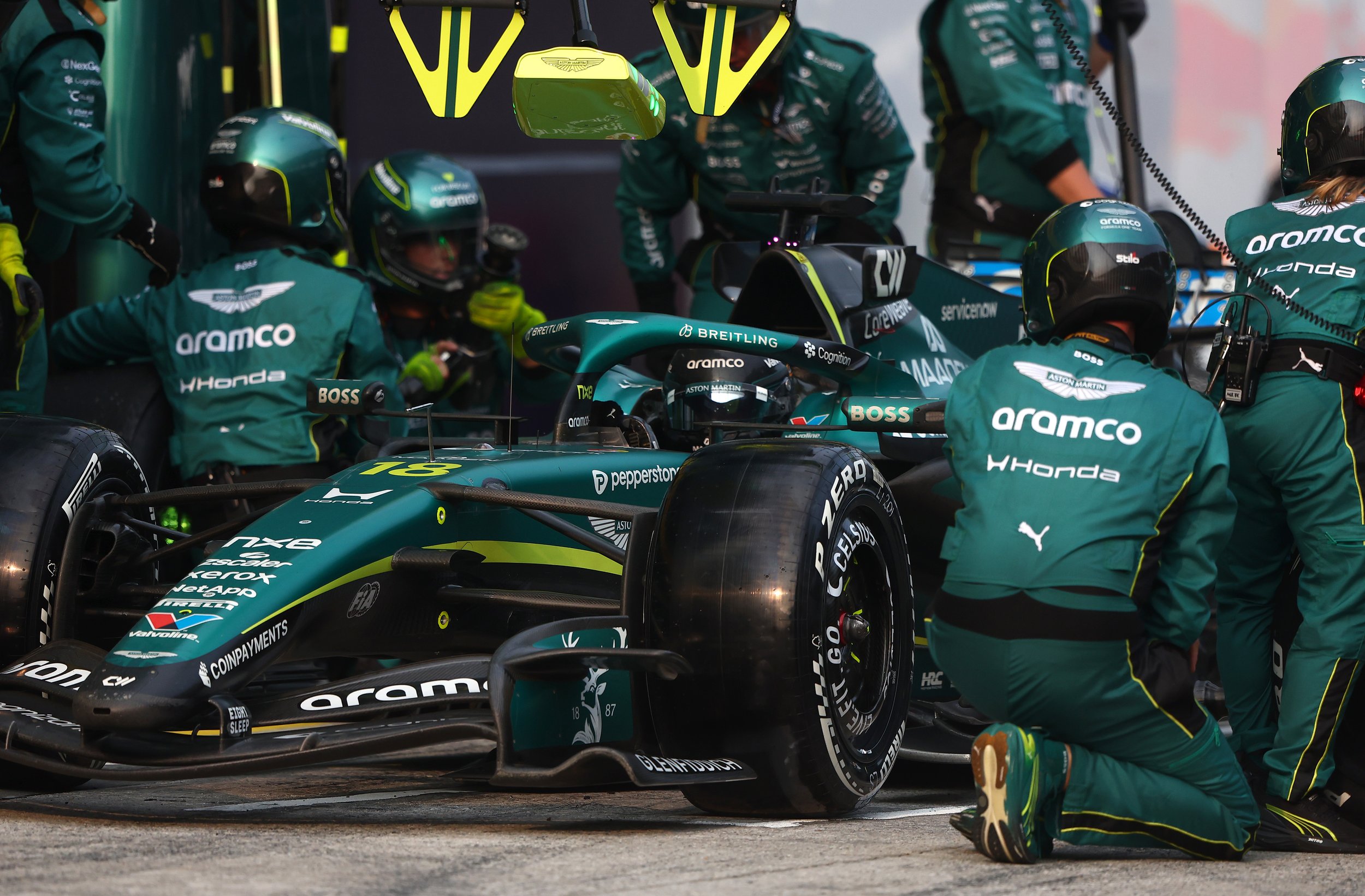 Team members working on a green Formula 1 race car in the garage during a race, with team uniforms and helmets visible.