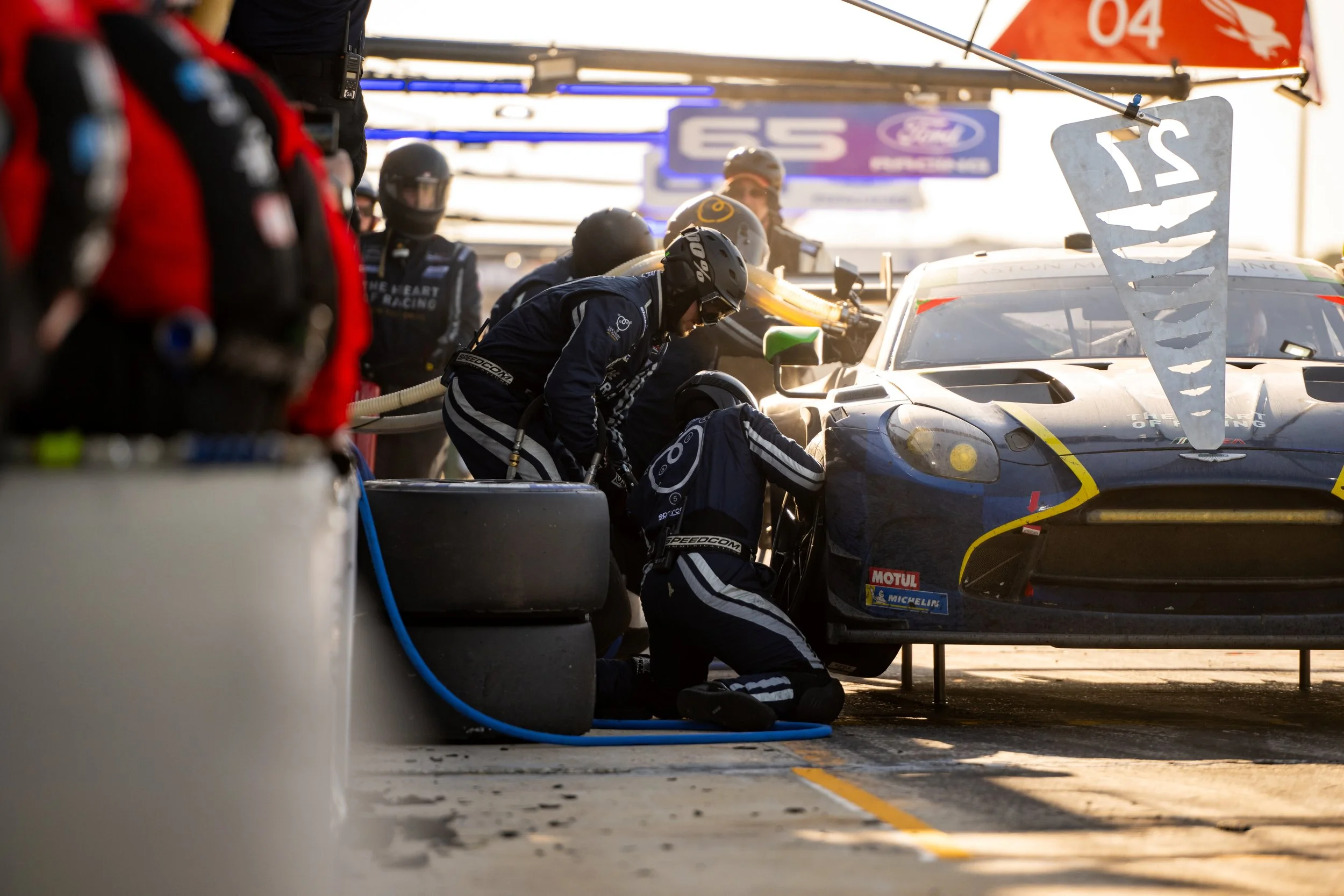 Pit crew members wearing helmets and racing suits working on a race car in a garage during a race event.