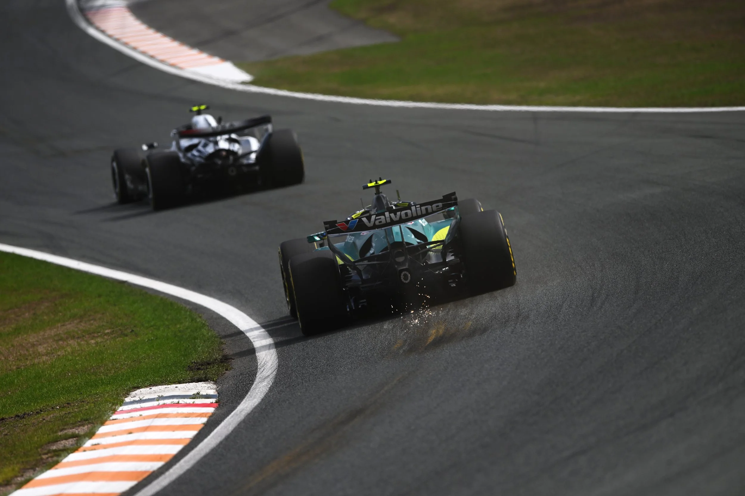 Two race cars on a racetrack, one black and the other turquoise, with the turquoise car leading around a curve, sparks flying from the rear of the turquoise car.