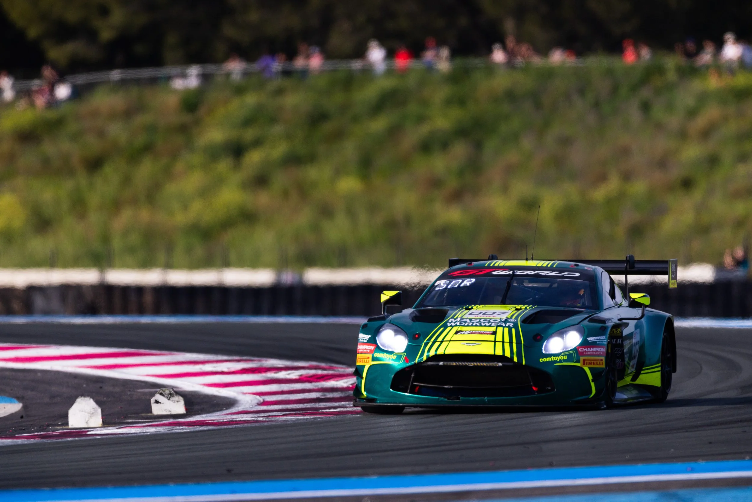 A green racing car with yellow and black accents navigating a turn on a race track, with a blurred background of spectators and trees.