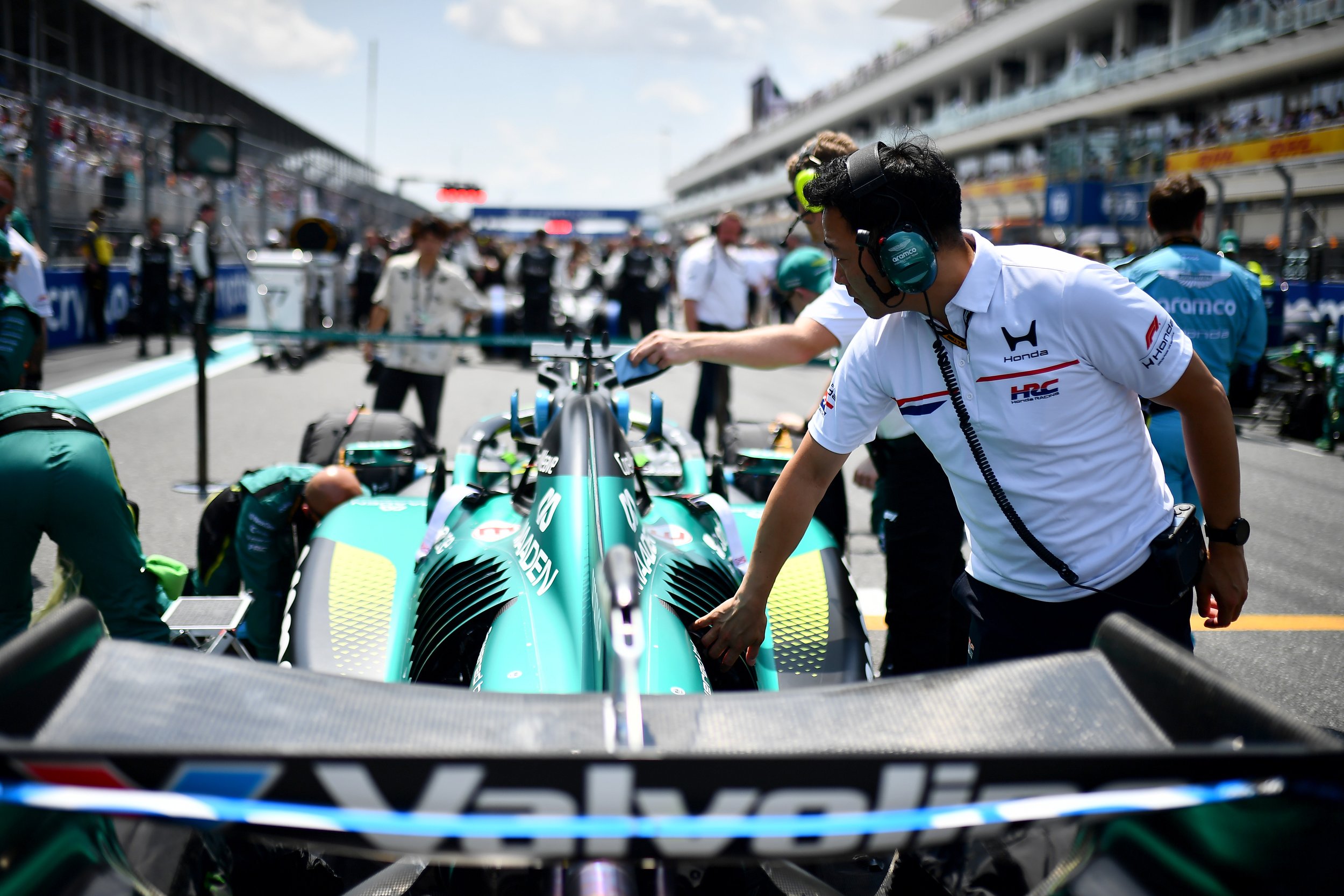 View from inside a racing car cockpit showing the front of a race car and team members working on it in a pit lane during a race event at a racing circuit.