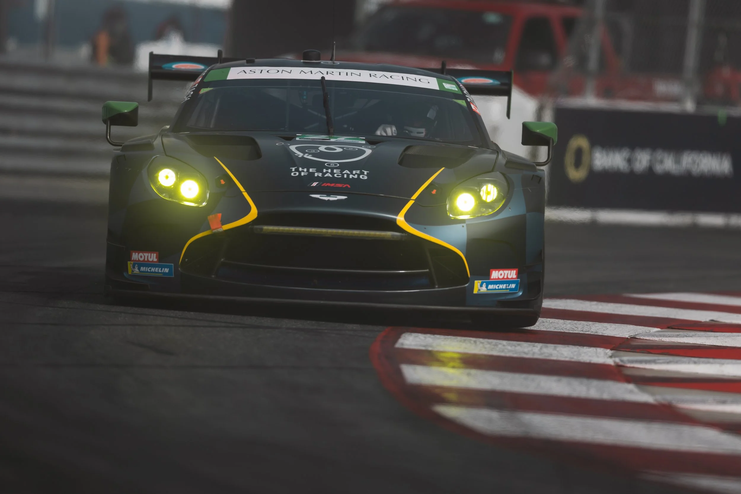 A Heart of Racing Aston Martin Vantage GT3 race car with yellow accents and bright yellow headlights on a racetrack during a race.
